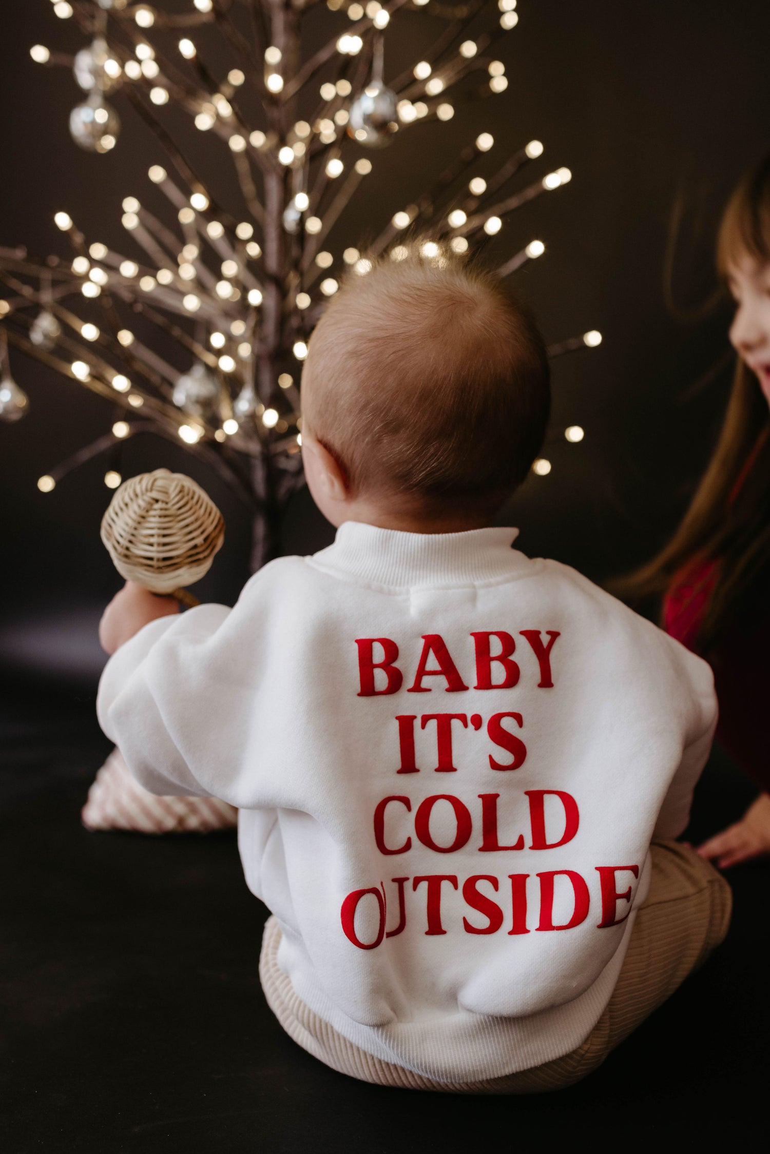A baby in a forever french baby "Child Sweatshirt | Baby It's Cold Outside" with bold red lettering sits in front of a decorative, illuminated tree, creating the perfect holiday season apparel scene.