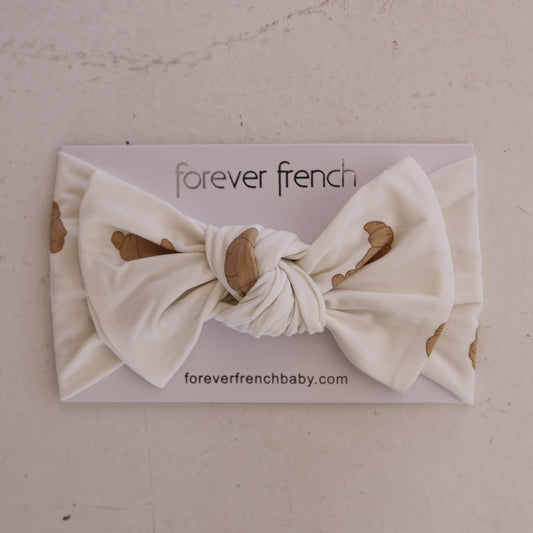 The Bamboo Head Wrap | Croissants from forever french baby features a white band with a large bow and croissant print, combining comfort and style for your little one. Displayed on a branded card, it's perfect for any occasion.