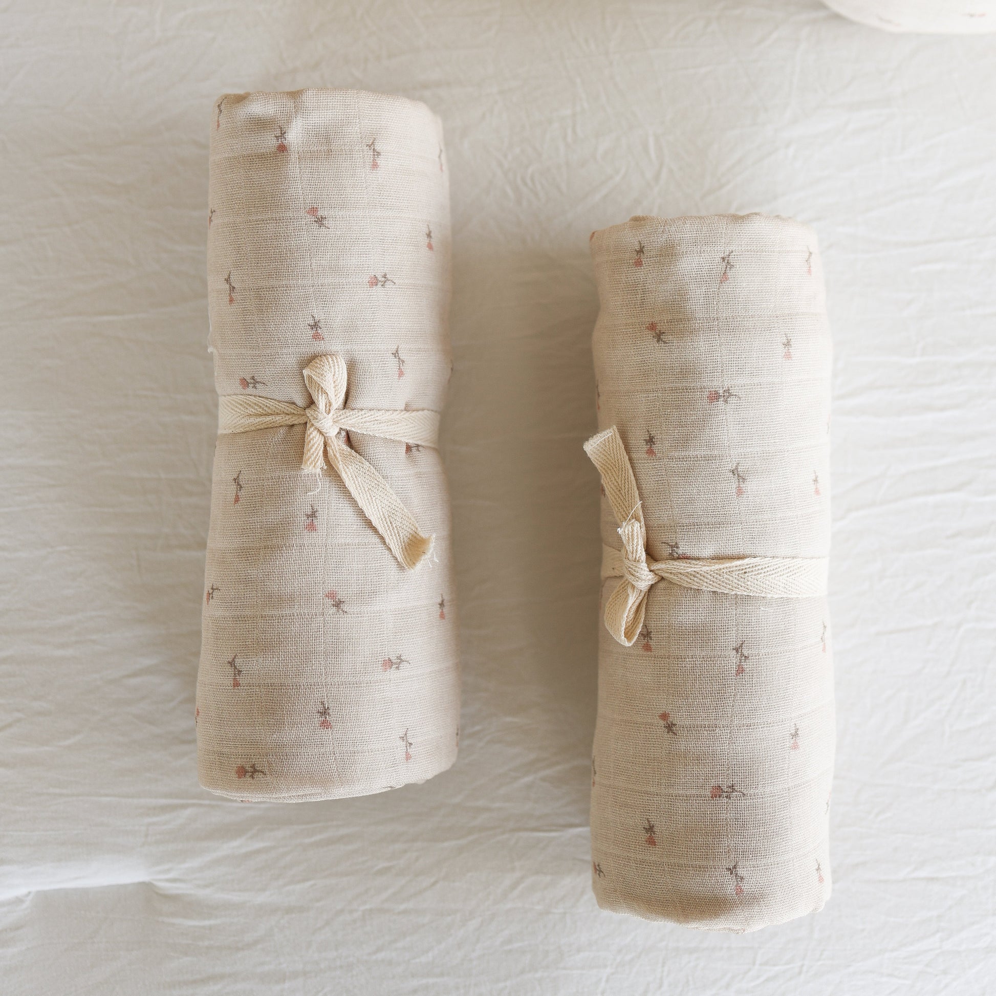 Two rolled-up forever french baby Muslin Swaddle | Lucy blankets in beige with delicate floral patterns, each tied with a ribbon, rest on a white textured surface.