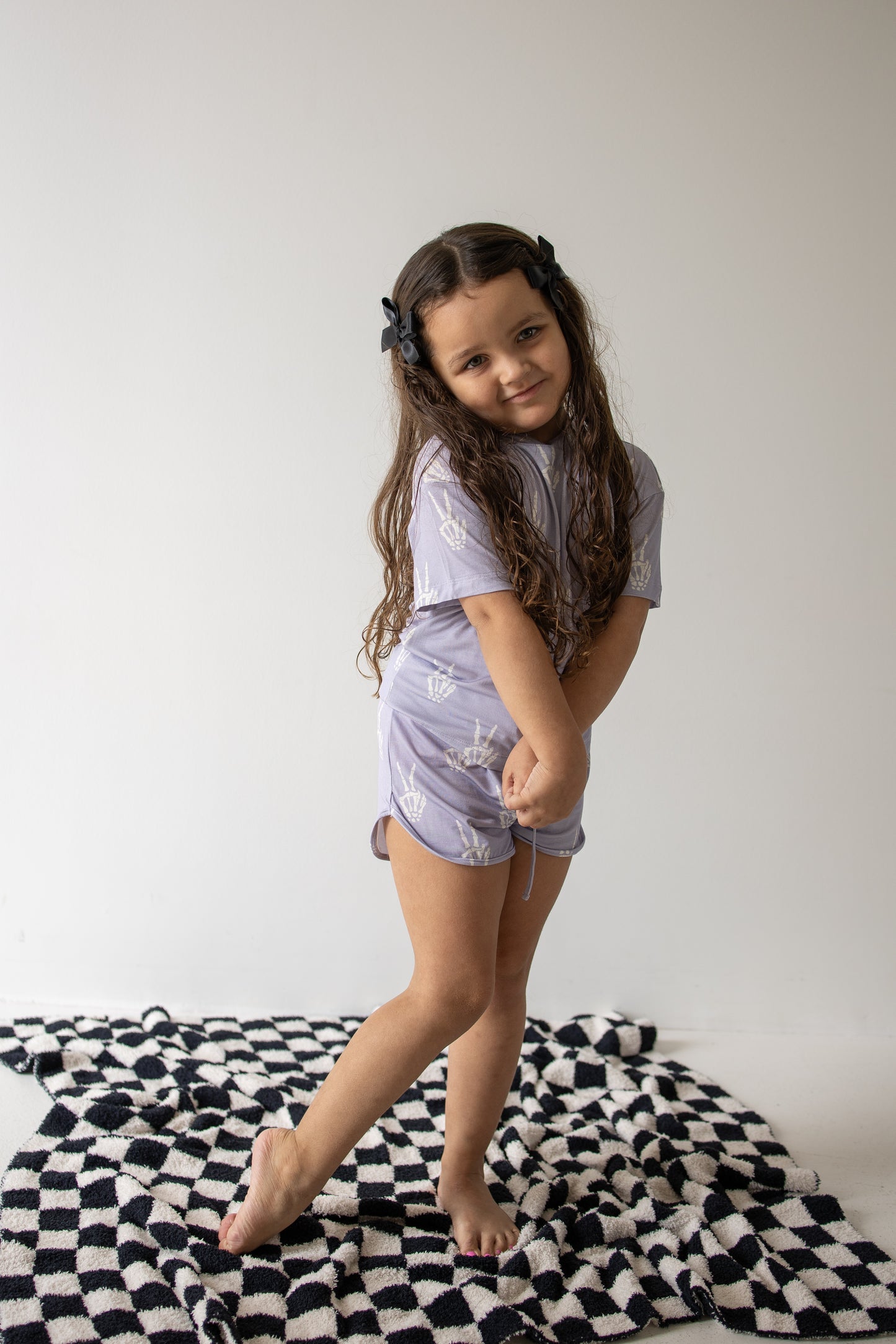 A young girl with long brown hair stands barefoot on a black and white patterned rug, smiling gently as she wears the forever french baby Girl’s Bamboo Short Set | Enchanted Skelly. She tilts her head and clasps her hands in front of her, enjoying the comfort of these hypo-allergenic kids clothes.