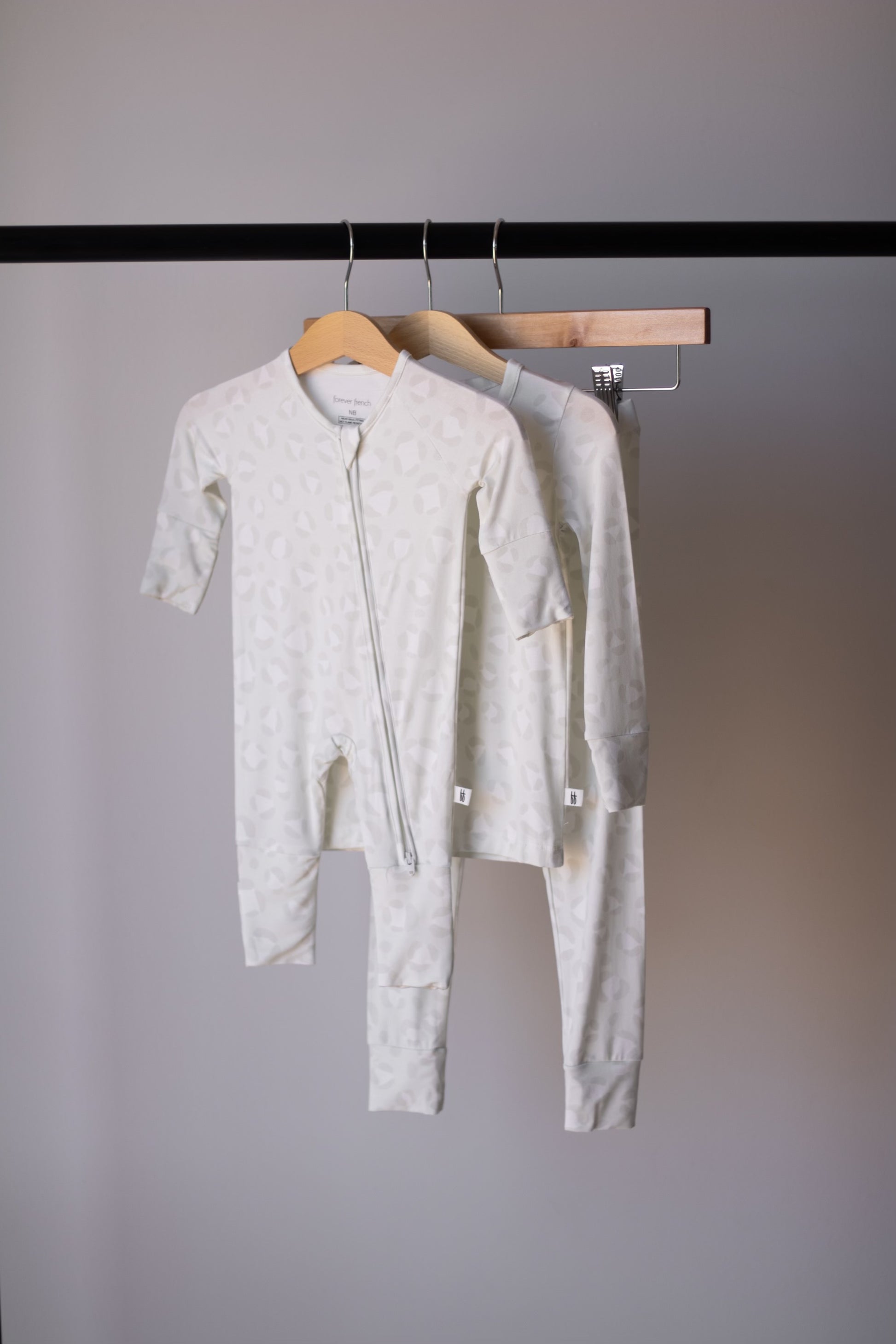 Two light-colored Bamboo Two Piece Pajamas | Meow by forever french baby, featuring subtle patterns and long sleeves for babies and toddlers, hang on wooden hangers from a black clothing rack against a plain gray background.