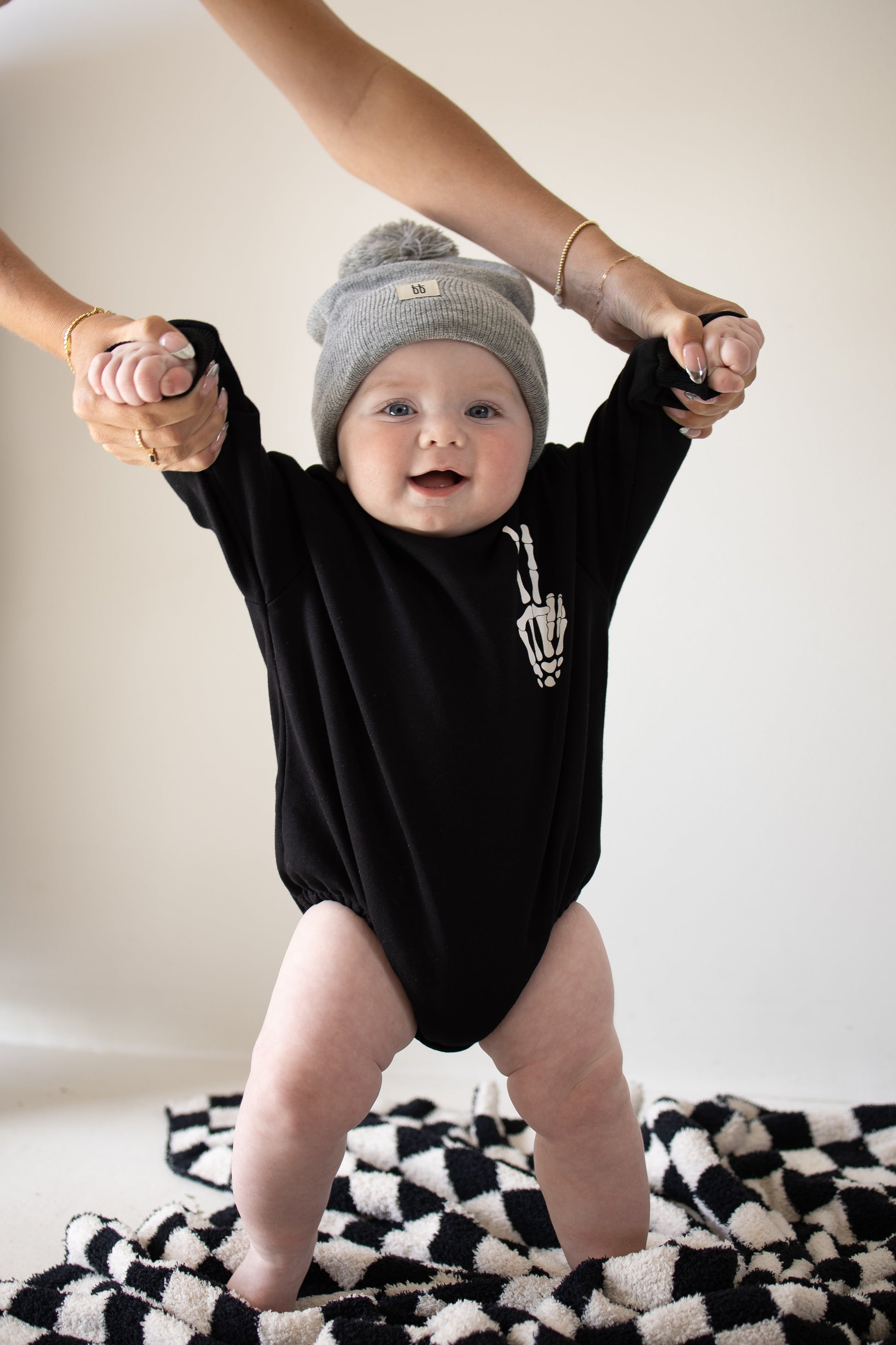 A smiling baby in a black “Sweatshirt Romper | Midnight Skelly” by forever french baby and a gray beanie is held up by an adult’s hands, standing on a black-and-white patterned blanket against a plain background. The cozy romper is part of the forever french baby collection and features the playful Midnight Skelly design.