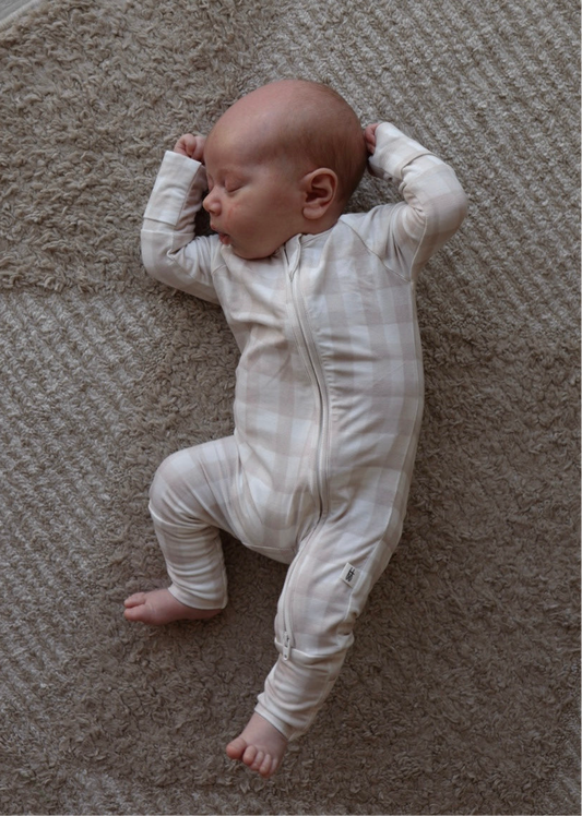 A newborn, dressed in forever french baby Bamboo Zip Pajamas | Gingham, sleeps with arms raised and legs relaxed on a textured beige carpet.