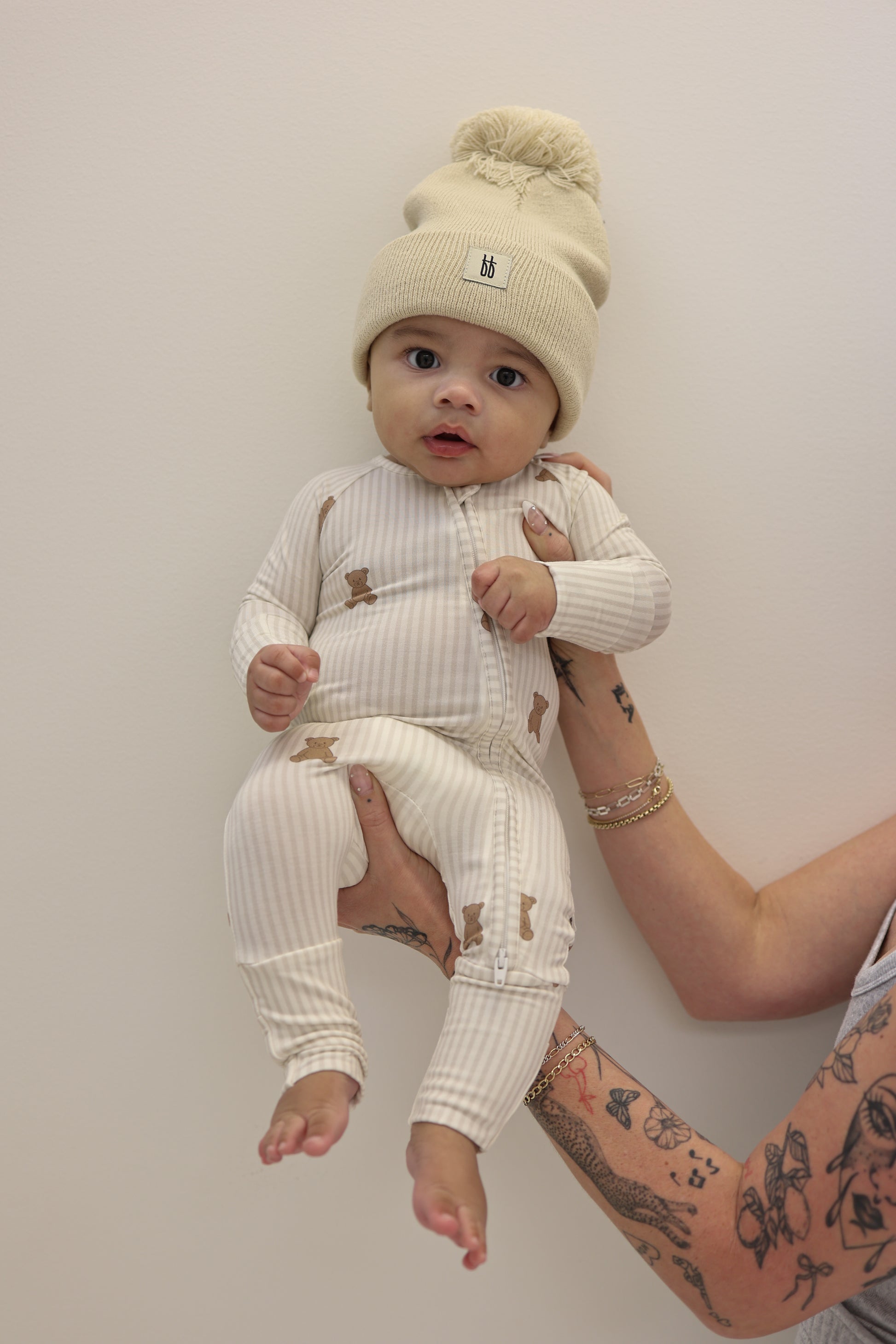 A person with tattooed arms holds a baby in a cream knit hat and forever french baby Bamboo Zip Pajamas | Teddy, against a plain light background.