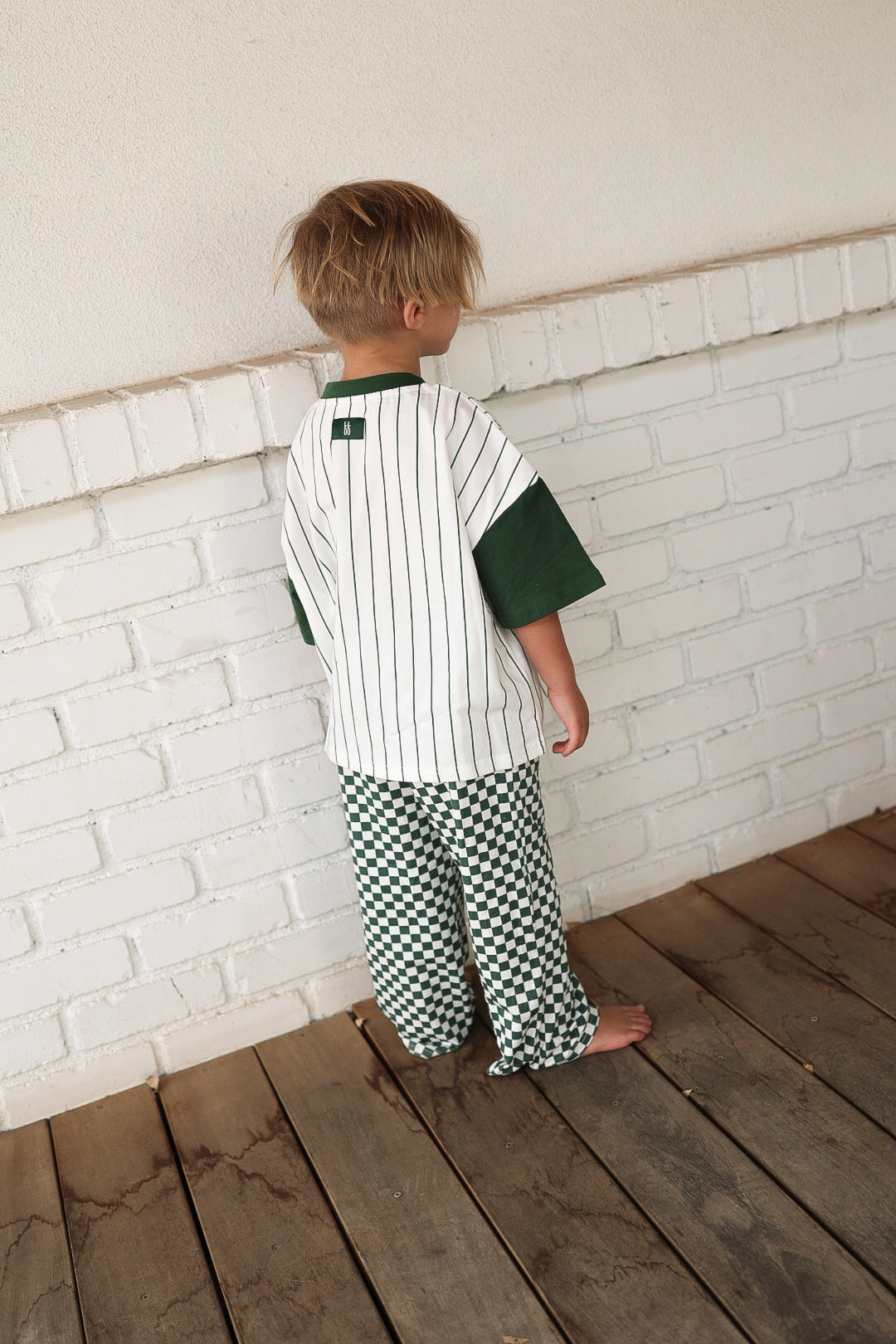 A young child stands barefoot on a wooden floor, facing a white brick wall, wearing the forever french baby T-Shirt & Pant Set | Lucky Micro Check: a white pinstriped shirt with green sleeves and green-and-white checkered wide-leg pants.