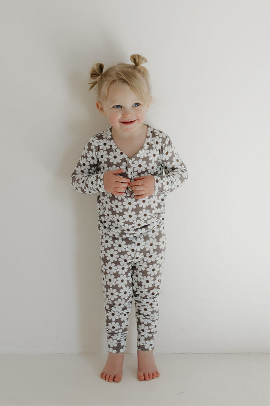 A young child with blonde pigtails smiles barefoot against a white wall, wearing forever french baby’s Bamboo Two Piece Pajamas | Darlin'—long-sleeve pajamas featuring a white daisy print on a gray background.