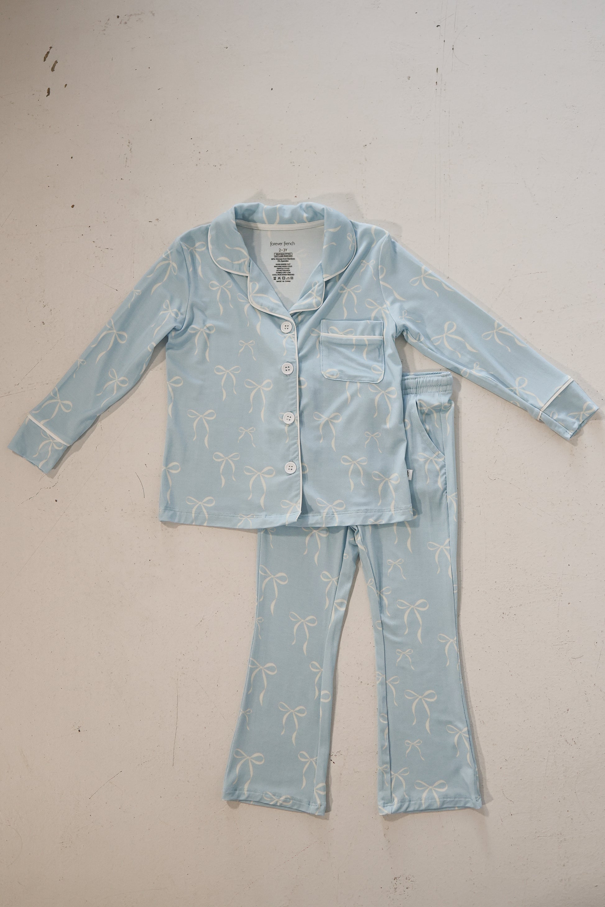 The Girl’s Bamboo Button Up Lounge Set | Blue Bows by forever french baby features a hypo-allergenic, light blue long-sleeve top with white bows and chest pocket, plus matching pants, displayed on a light-colored surface.