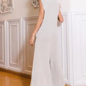 A person wearing the Bucketlist Wide Leg Button-Up Loose Fit Solid Sweater Jumpsuit, a sleeveless and light-colored outfit, stands with their back to the camera in a room with wainscoting and framed wall art. The jumpsuit features wide-leg pants and has a texture that appears soft and comfortable.