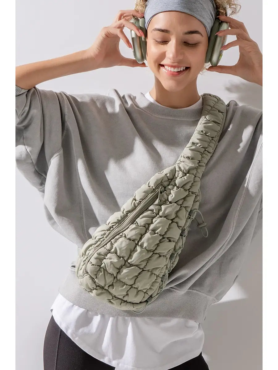 A person is smiling with eyes closed, wearing a grey headband, grey oversized sweatshirt, and headphones. They have an urban daizy Quilted Sling Bag in light green slung over their shoulder – a stylish accessory perfect for everyday use. The background is plain and well-lit.