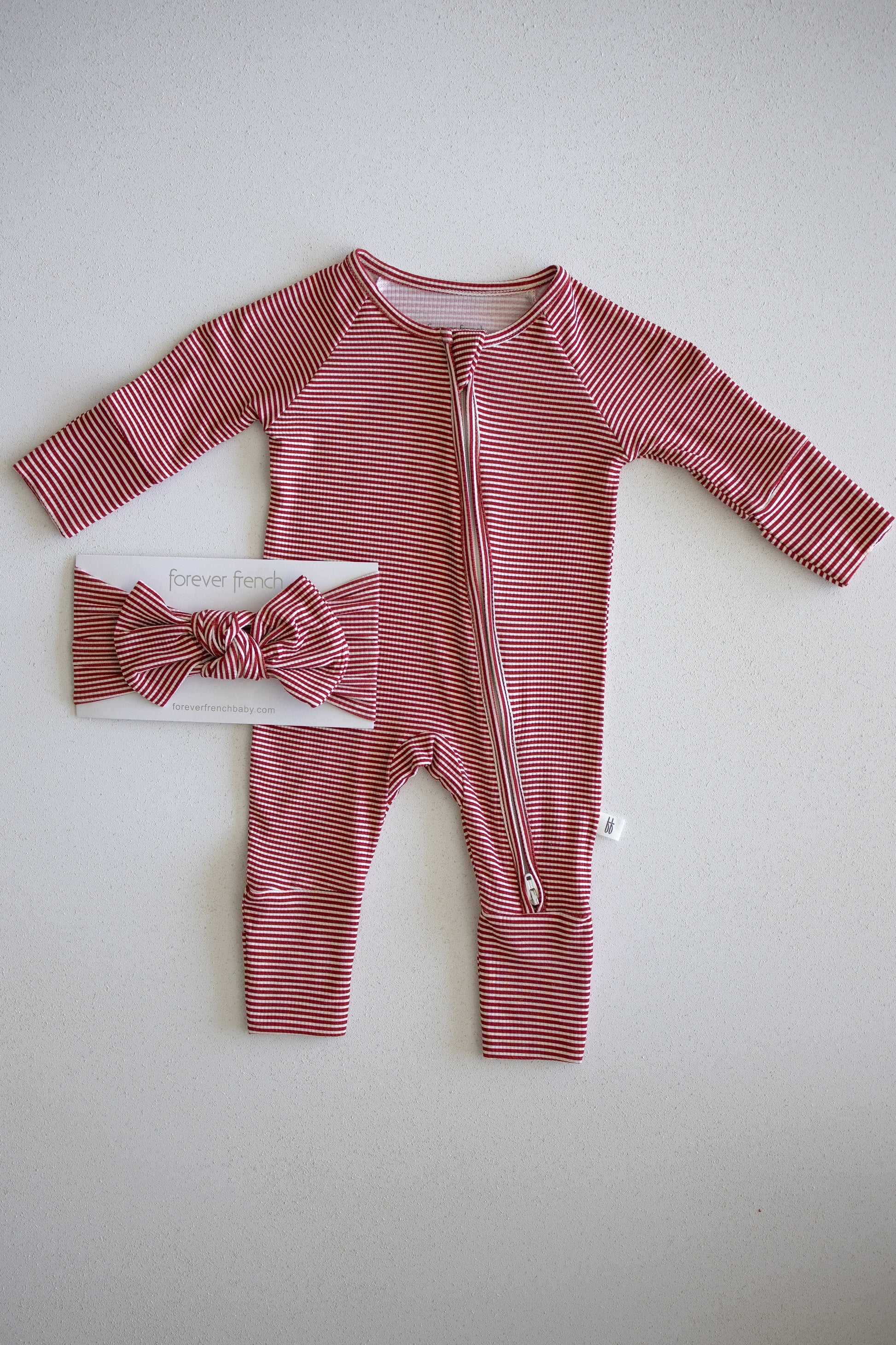 Sure! Here’s a rewritten sentence using your product data: A red and white peppermint stripe baby onesie with a diagonal zipper is displayed next to the forever french baby Bamboo Head Wrap | Peppermint Stripe Ribbed on a white background.
