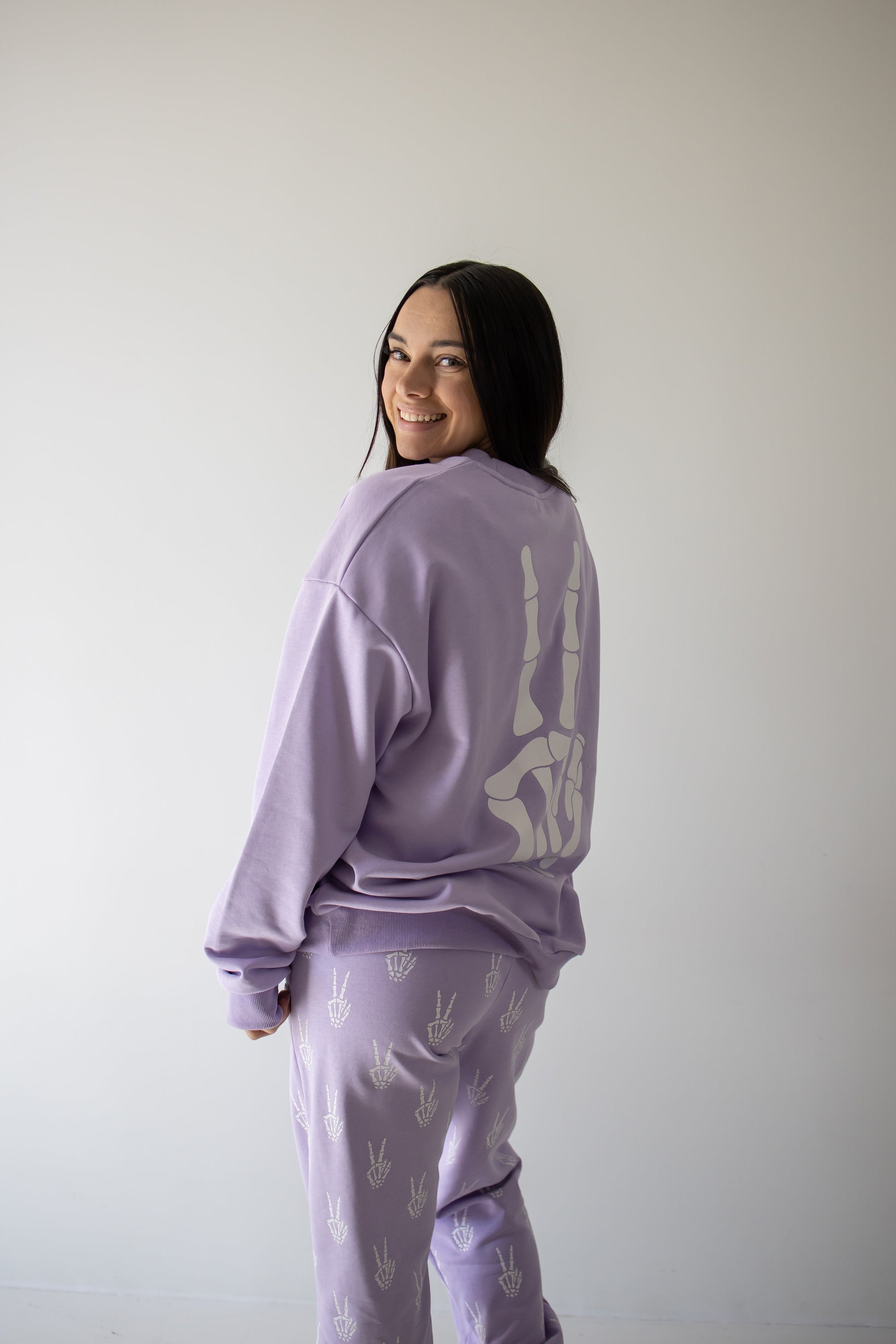 A woman smiles at the camera, standing against a plain light-colored wall in the forever french baby Women’s Sweat Set | Enchanted Skelly—a cozy lavender set adorned with white hand symbols, making it the perfect choice for a spooky season outfit.