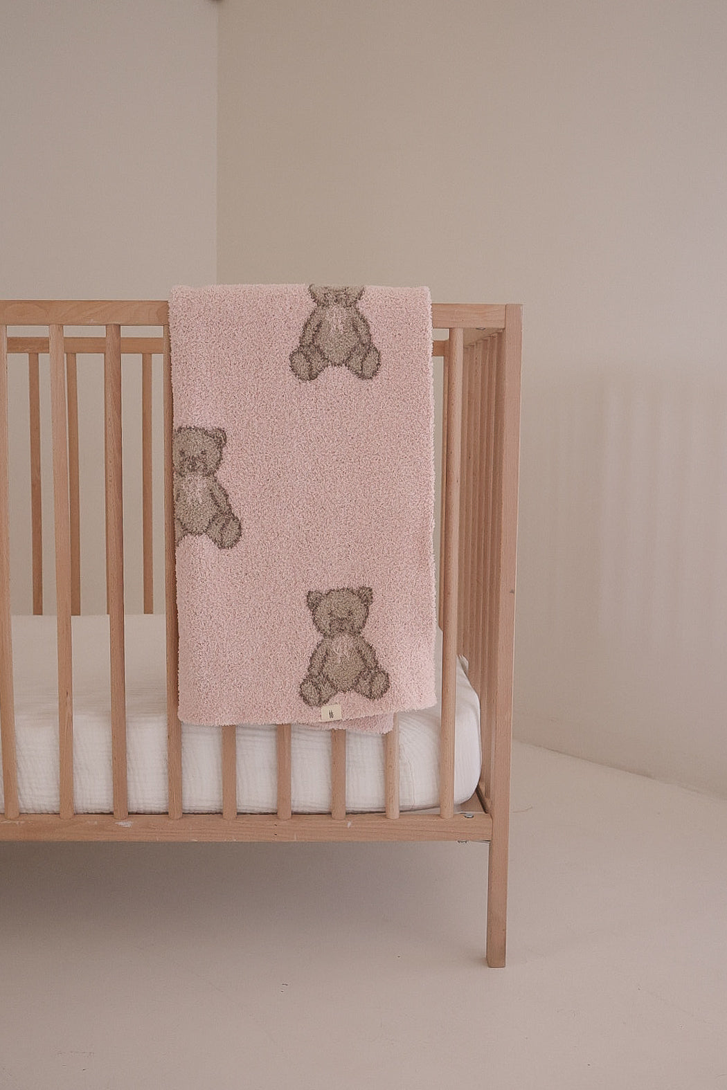 A light wooden crib with a white mattress stands in a minimal room. Draped over the rail is the forever french baby Plush Blanket | Lottie, an Oeko-Tex certified soft pink blanket featuring teddy bear designs.