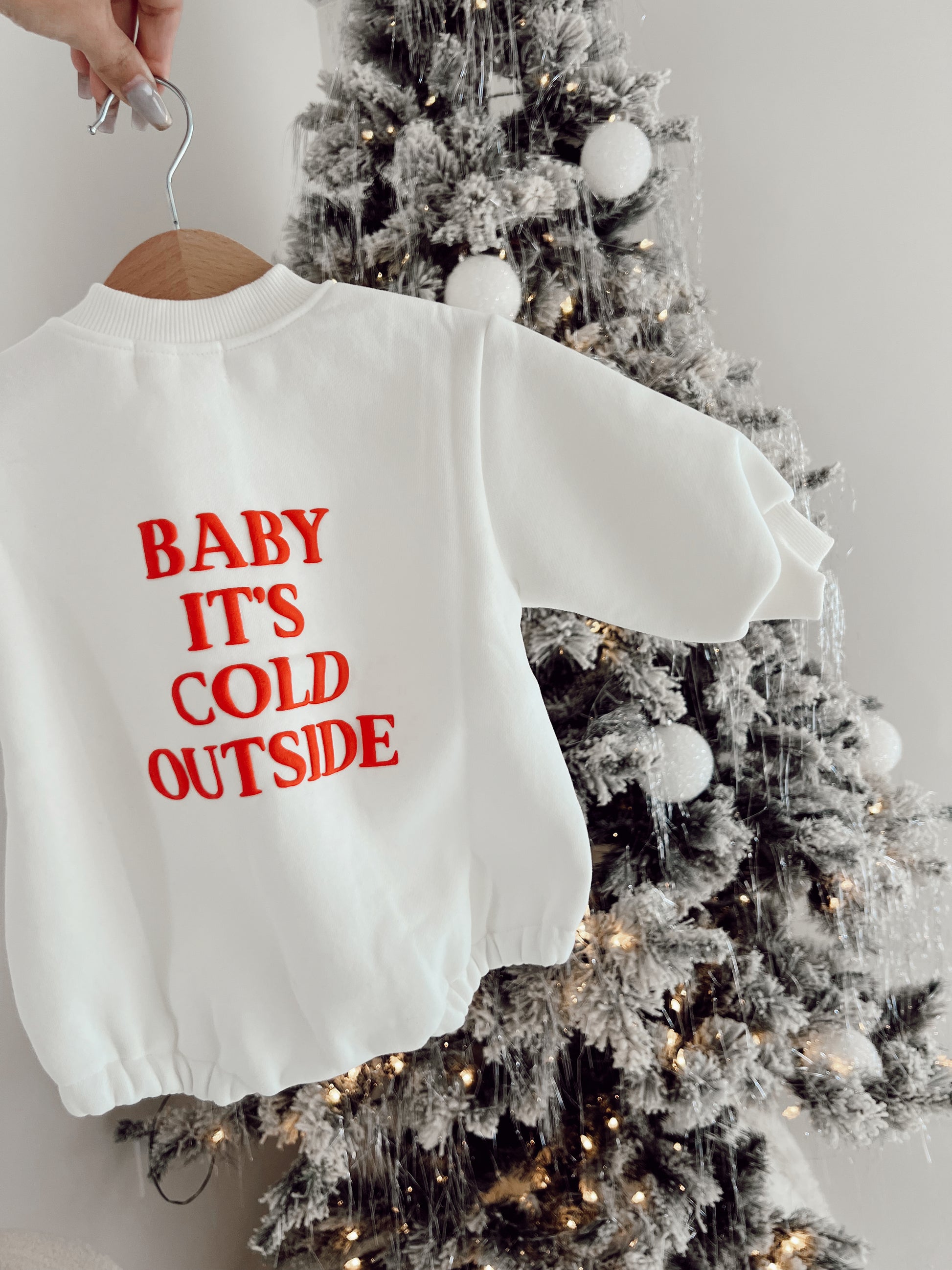 A hand holds a cozy oversized romper from forever french baby, featuring red text reading "Baby It's Cold Outside," against a snow-dusted Christmas tree adorned with white ornaments and twinkling lights—a perfect choice for chilly weather days.