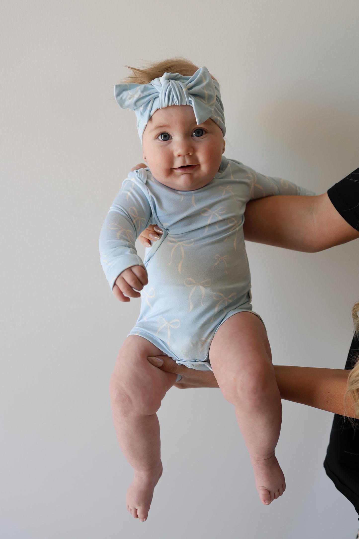 A baby in a light blue Long Sleeve Bamboo Snapsuit | Blue Bows by forever french baby, with a matching headband, is held up by an adult against a white wall, looking at the camera with a slight smile.