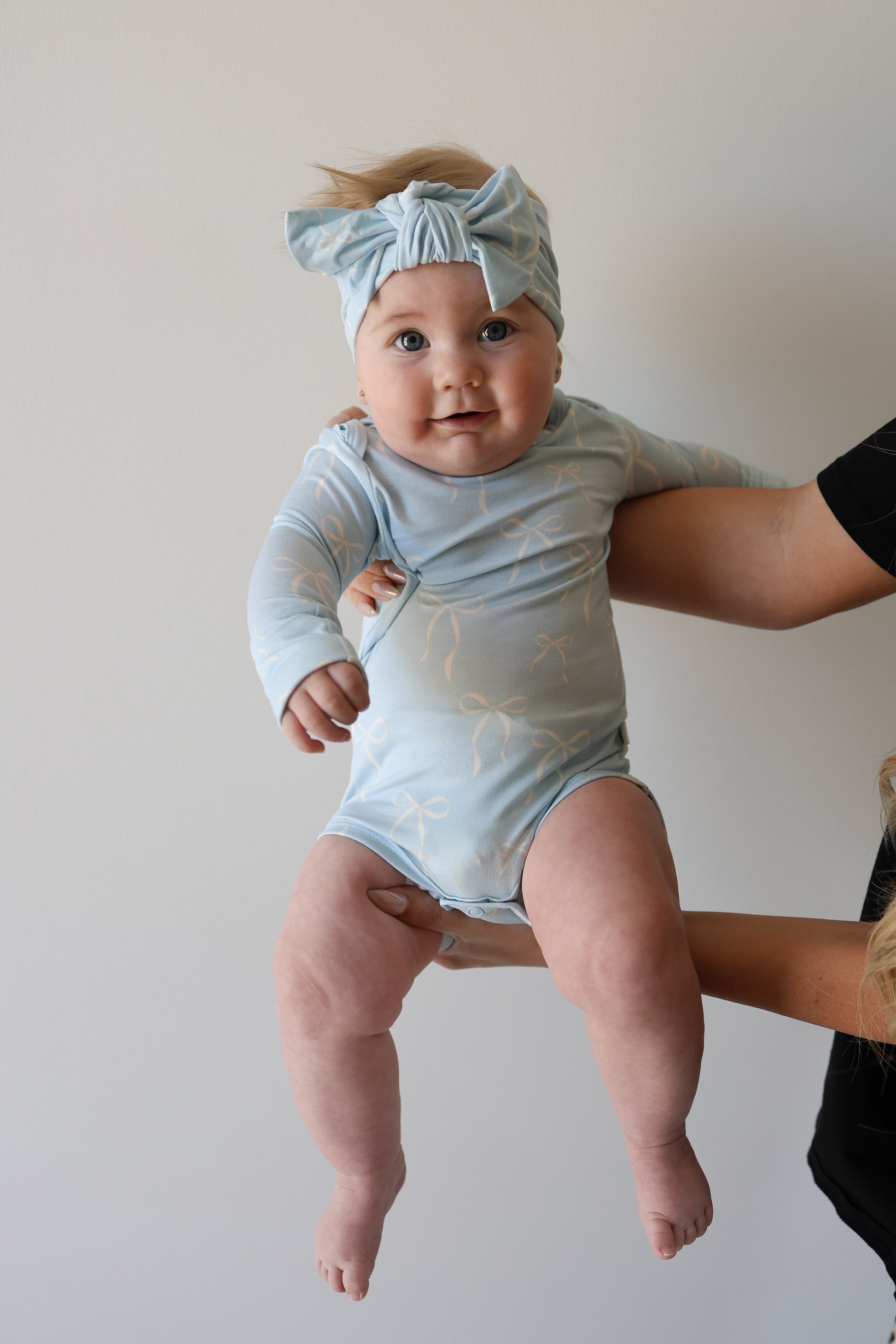 A baby in a light blue Long Sleeve Bamboo Snapsuit | Blue Bows by forever french baby, with a matching headband, is held up by an adult against a white wall, looking at the camera with a slight smile.