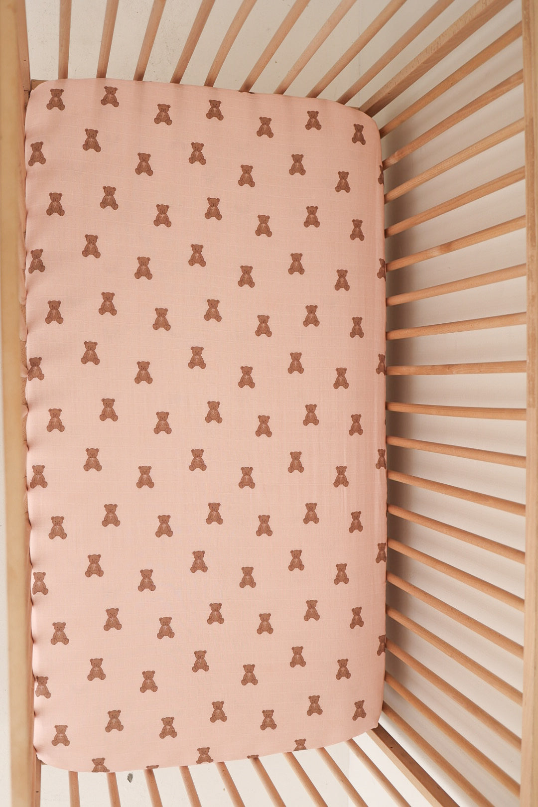 A top-down view of the forever french baby Muslin Crib Sheet | Pink Lottie, featuring a pink background with a repeating small brown teddy bear pattern—ideal for cozy and stylish baby bedding.