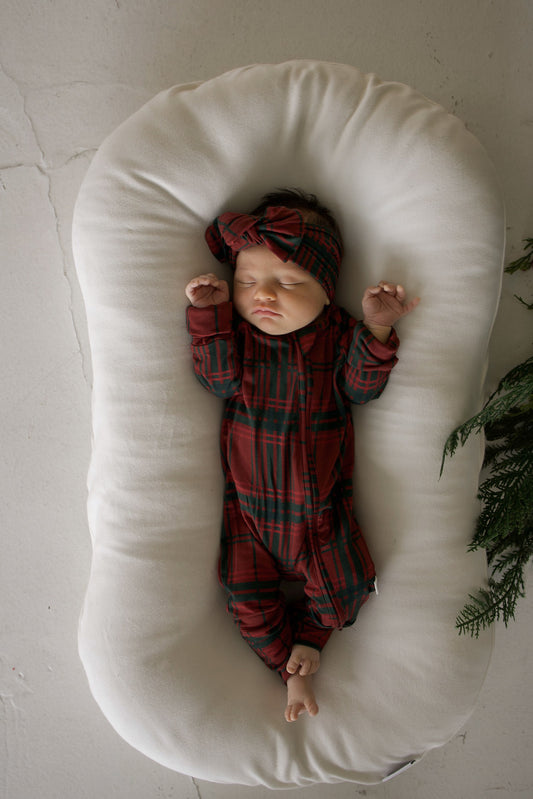 A newborn sleeps in Bamboo Zip Pajamas | Christmas Plaid and a matching headband, lying in a white cushioned lounger.