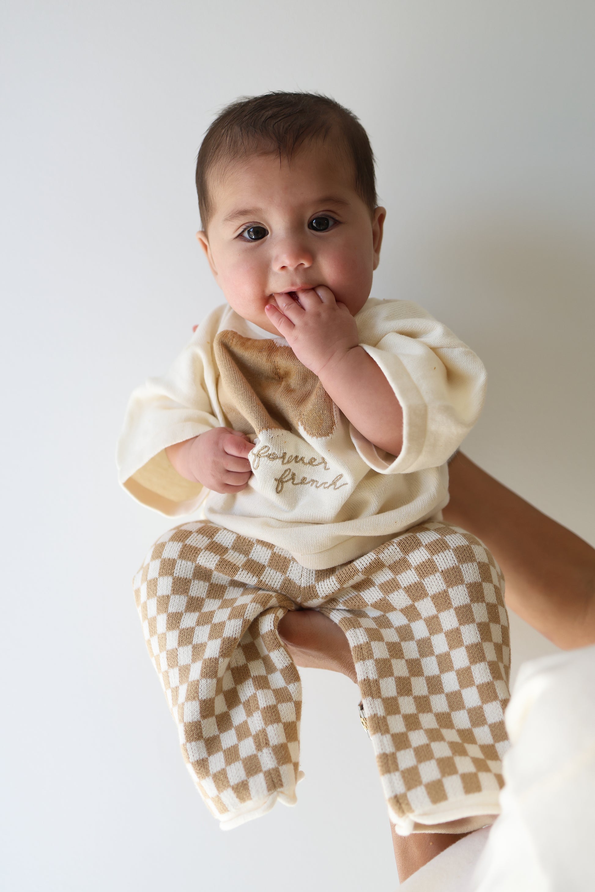 A baby with dark hair sits upright, wearing the forever french baby Knit Pant Set | Croissants—featuring a cream shirt and beige checkered pants—with fingers in their mouth and an adult’s hand supporting them against a plain white background.