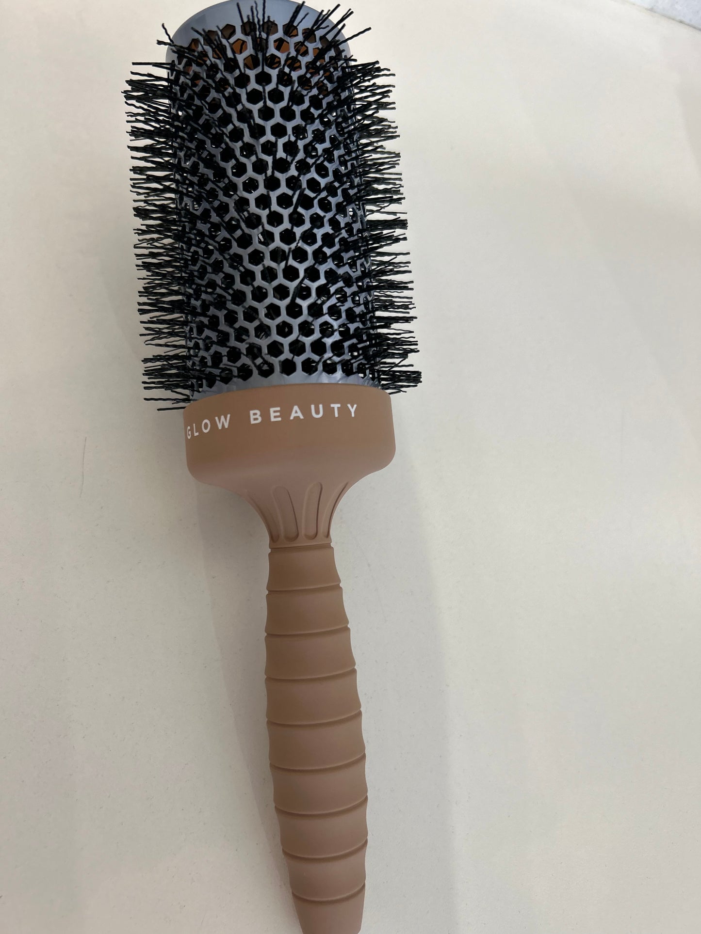 A round Glow Beauty Hair Brush from forever french baby, featuring black metal bristles, a grey barrel with the text "GLOW BEAUTY," and a light brown handle with a ribbed grip, lies on a white surface.