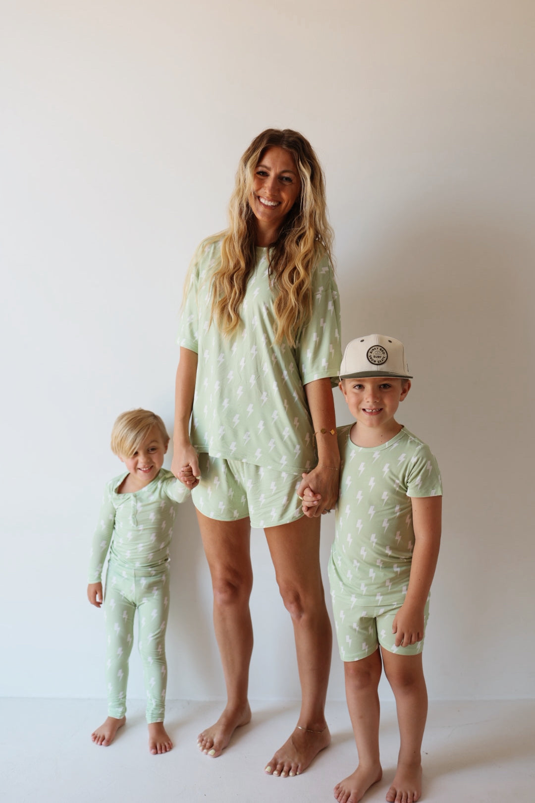 A woman and two young children stand barefoot against a white background, smiling and holding hands, all wearing the forever french baby Bamboo Short Set | Flash—matching light green bamboo pajamas with a white lightning bolt pattern. The older child wears a white cap.