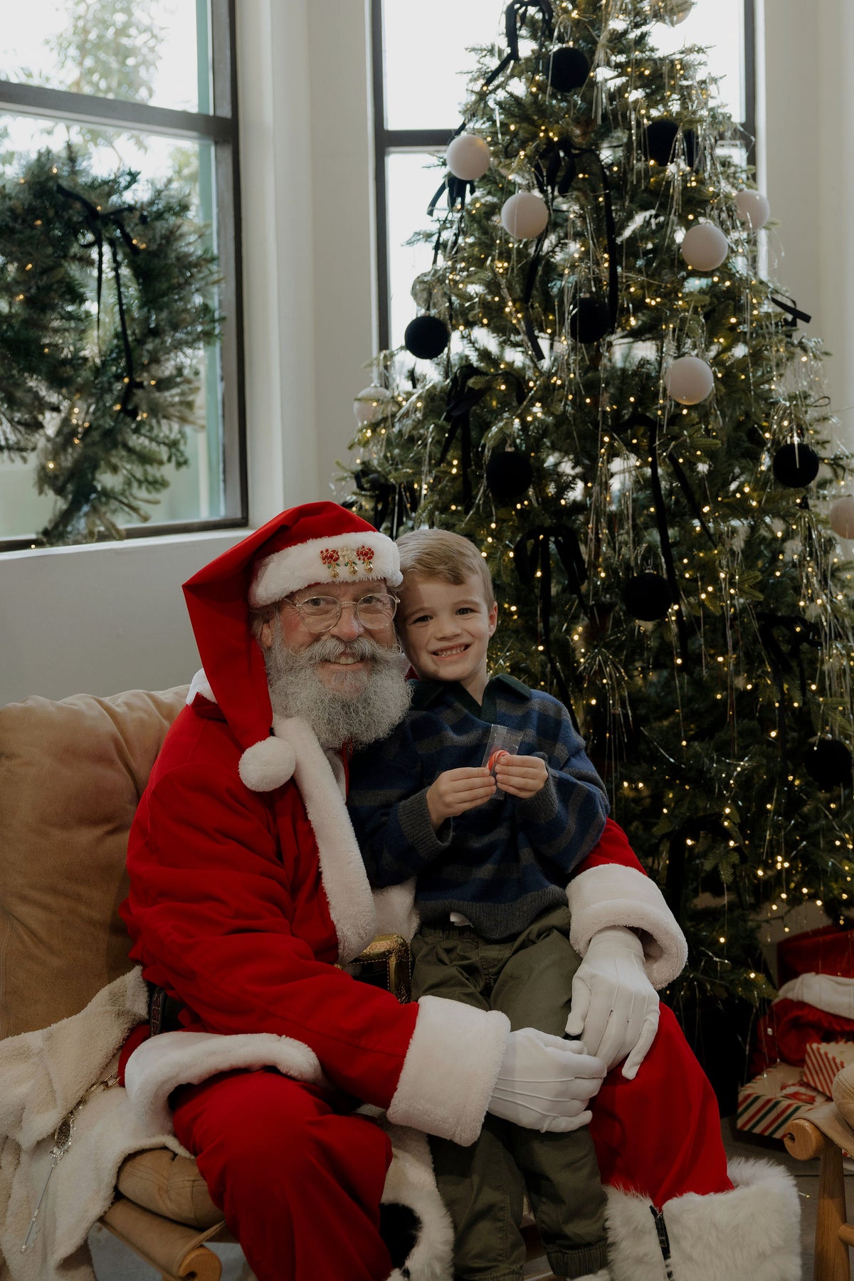 A young boy sits on Santa’s lap in front of a decorated Christmas tree for his Santa Photos | Forever French Baby x Emily Schuman Photography.