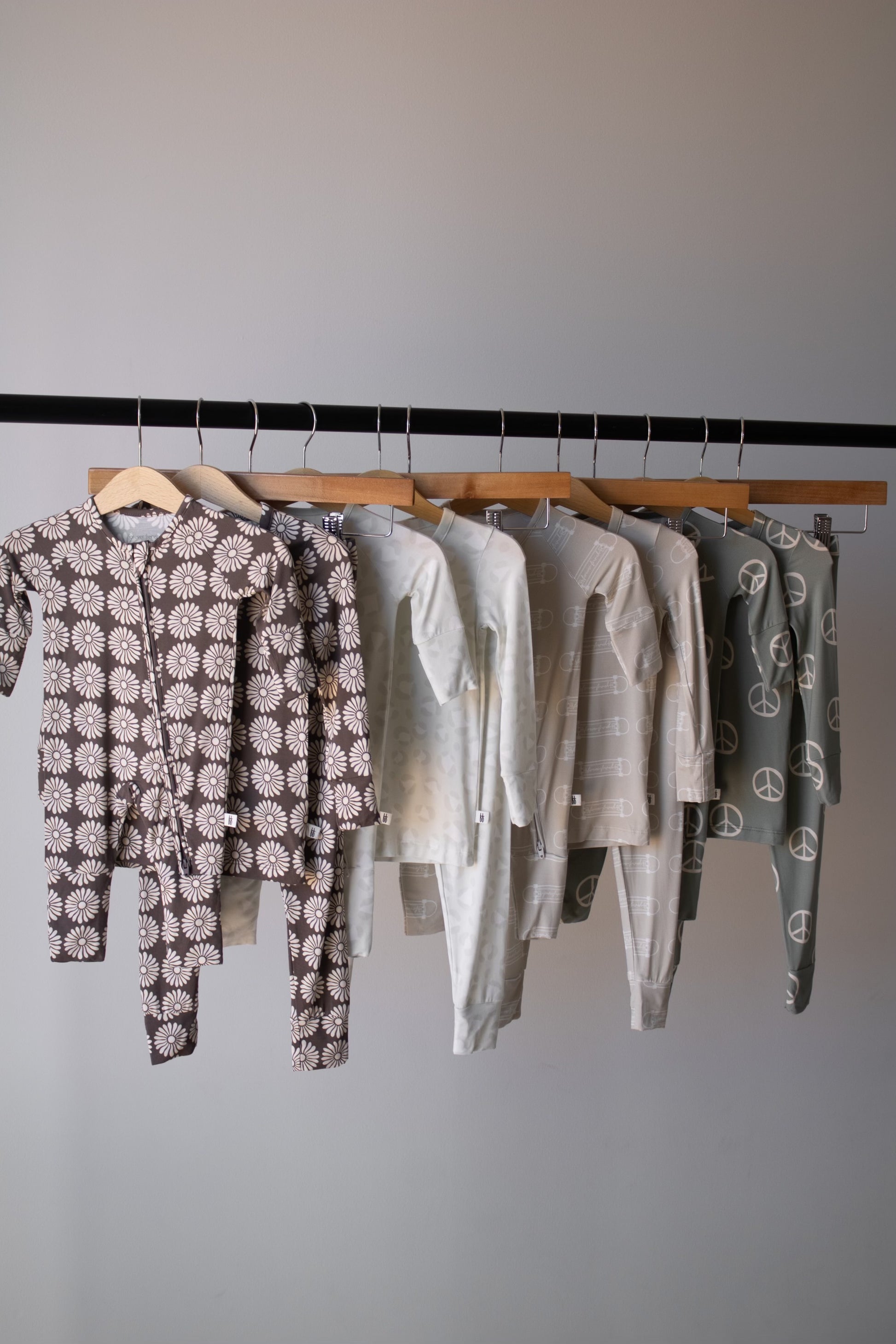 Six sets of forever french baby Bamboo Two Piece Pajamas | Sk8er Boy in neutral tones hang on a rack, each featuring breathable fabric and playful patterns like daisies, stars, stripes, and peace signs.