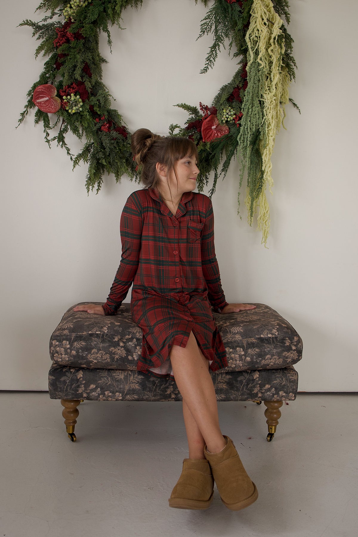 A girl sits on a floral ottoman wearing a long-sleeved Bamboo Sleeping Dress | Christmas Plaid.