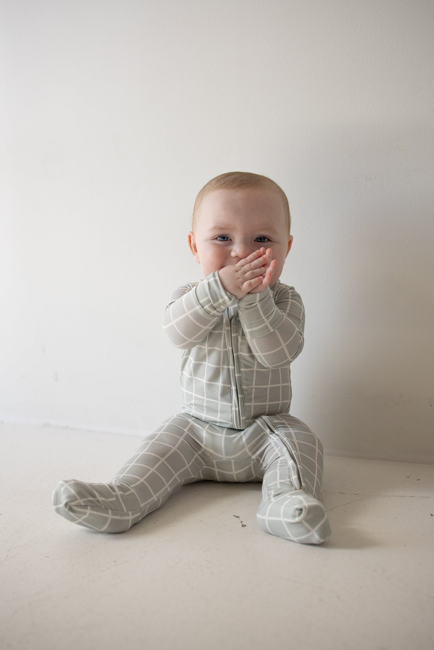 A baby in the forever french baby Bamboo Zip Pajamas | Foggy Grid, featuring a soft gray checkered design, sits on the floor against a white wall, clapping their hands and smiling warmly at the camera.