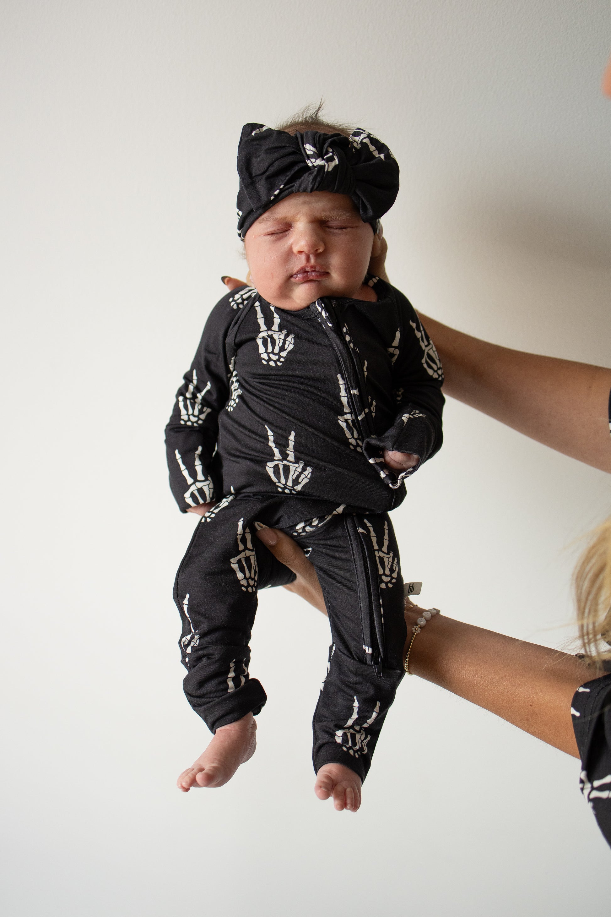 A baby dressed in forever french baby’s Bamboo Zip Pajamas | Midnight Skelly, featuring soft black bamboo viscose with white skeleton hand prints and a matching headband, is held up against a plain white background—highlighting these stylish, hypo-allergenic pajamas.