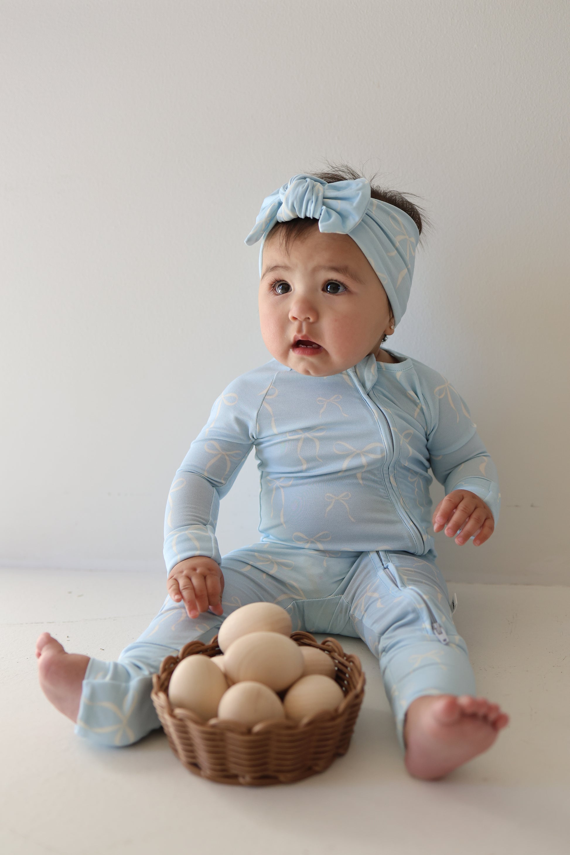 A baby in forever french baby’s Bamboo Zip Pajamas | Blue Bows sits on the floor beside a small wicker basket of brown eggs, wearing a matching headband, all against a plain white background.