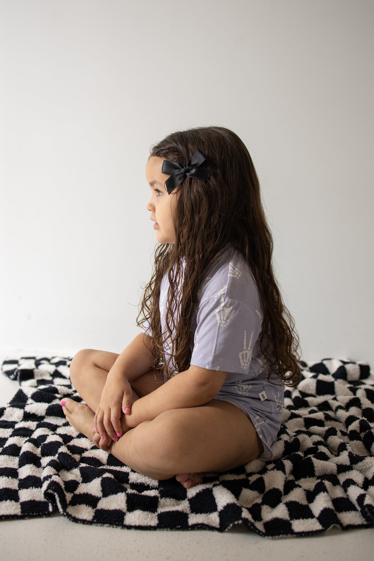 A young girl with long brown hair and a black bow sits cross-legged on a black-and-white checkered blanket, facing left against a plain light background, dressed in the forever french baby Girl’s Bamboo Short Set | Enchanted Skelly—a cozy, hypo-allergenic outfit that highlights the comfort and softness of bamboo kids clothes.