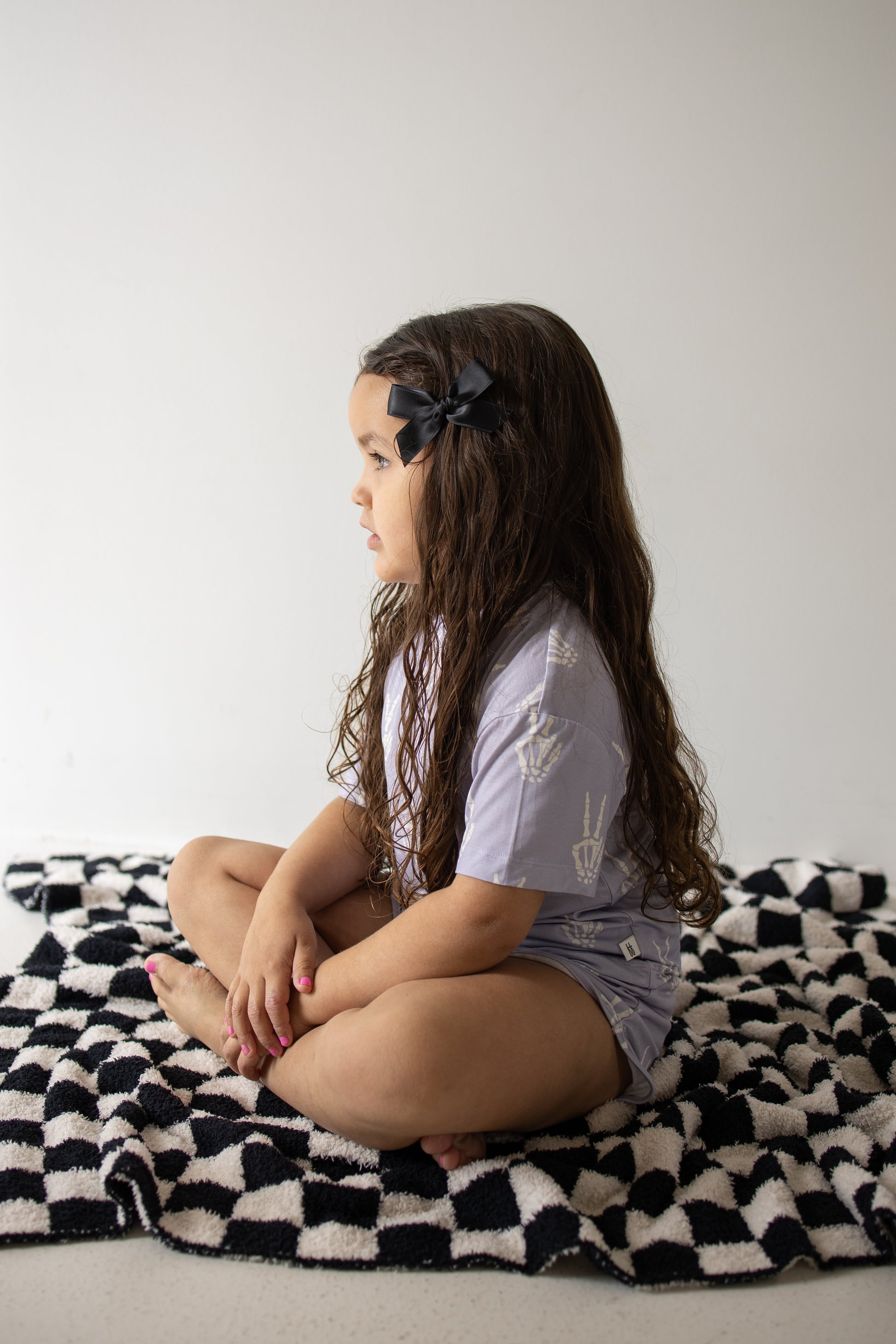 A young girl with long brown hair and a black bow sits cross-legged on a black-and-white checkered blanket, facing left against a plain light background, dressed in the forever french baby Girl’s Bamboo Short Set | Enchanted Skelly—a cozy, hypo-allergenic outfit that highlights the comfort and softness of bamboo kids clothes.
