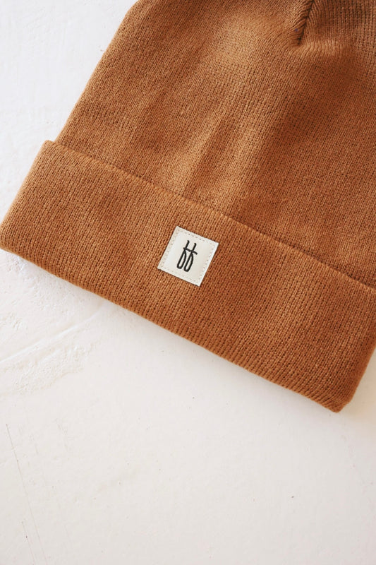 A close-up image showcases the ff Knit Beanie | Adult by forever french baby, featuring a brown knit design with ribbed details and a folded brim. The small rectangular white label on the brim displays a simple black design. Made of breathable yarn, the beanie is placed on a light, textured surface.