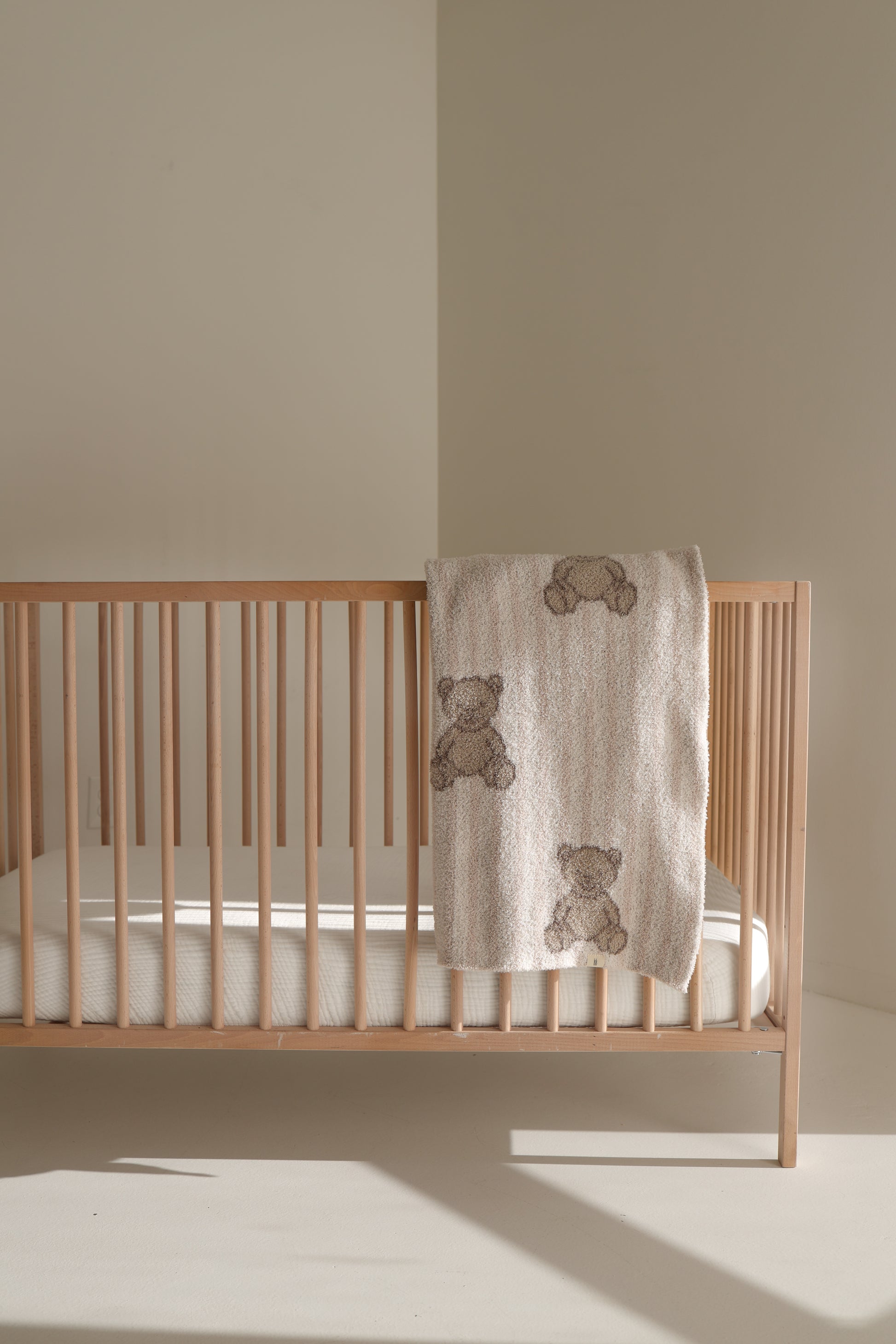 A wooden baby crib with a white mattress stands in a softly lit room. A forever french baby Plush Blanket | Teddy, Oeko-Tex certified and decorated with teddy bear designs, is draped over the side of the crib.