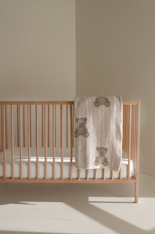 A wooden baby crib with a white mattress stands in a softly lit room. A forever french baby Plush Blanket | Teddy, Oeko-Tex certified and decorated with teddy bear designs, is draped over the side of the crib.