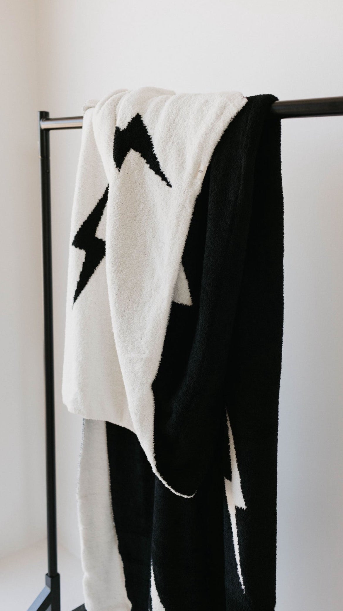 A "Plush Blanket | Black & White Lightning Bolt" by forever french baby, featuring a striking lightning bolt design, is elegantly draped over a black metal clothing rack. Crafted from microfiber feather yarn, the blanket primarily showcases white with black lightning bolts and black with white lightning bolts, creating a captivating contrast. For added peace of mind, it is OEKO Standard 100 certified.