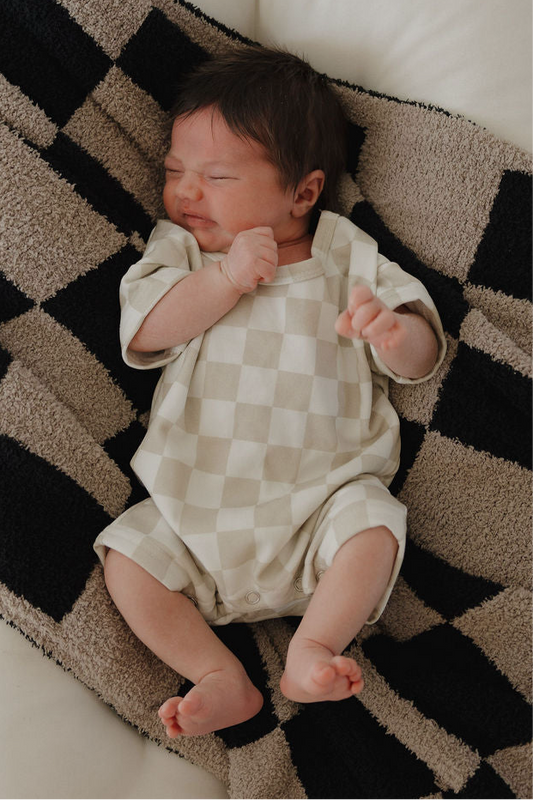 A newborn with dark hair sleeps on a patterned blanket, wearing the forever french baby French Terry Romper | Checkerboard—an adorable light checkered outfit with snap closures and cozy style.