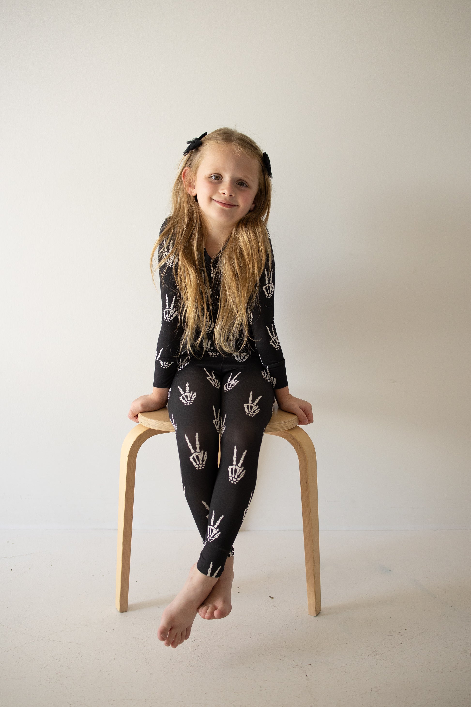 A young girl with long blonde hair sits barefoot on a wooden stool, wearing forever french baby’s Bamboo Two Piece Pajamas | Midnight Skelly, featuring white skeleton hand patterns, against a plain light-colored wall.
