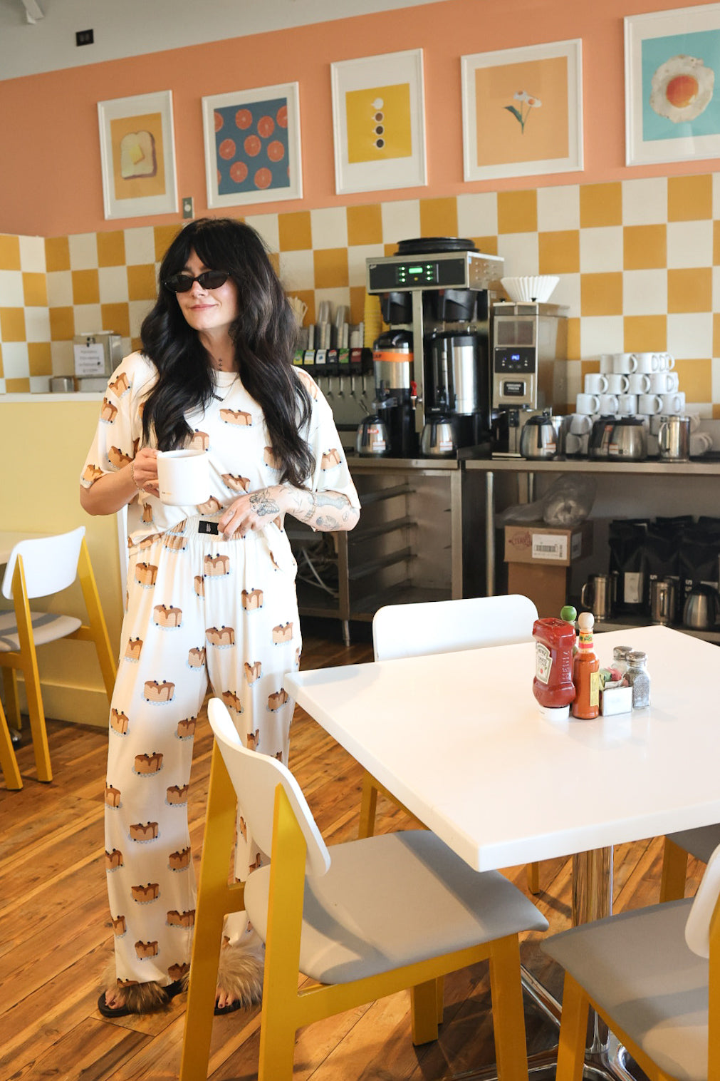 A woman with long dark hair and sunglasses stands in a bright cafe holding a mug, wearing the Women's Boxy Tee Lounge Set | Sunday Stack by forever french baby. The cafe has yellow checkered walls, framed art, and a coffee machine.