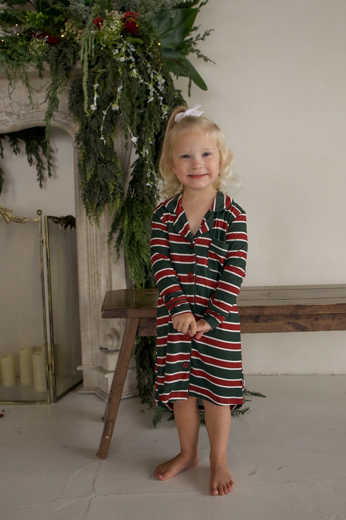 A young girl wearing a Bamboo Sleeping Dress | Holiday Stripe, a long-sleeved nightshirt with red, white, and green horizontal stripes.