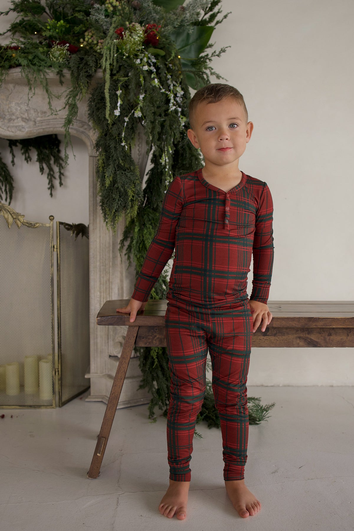 A young boy wears a long-sleeved, henley-neckline Bamboo Two Piece Pajamas | Christmas Plaid set while standing by a wooden bench.