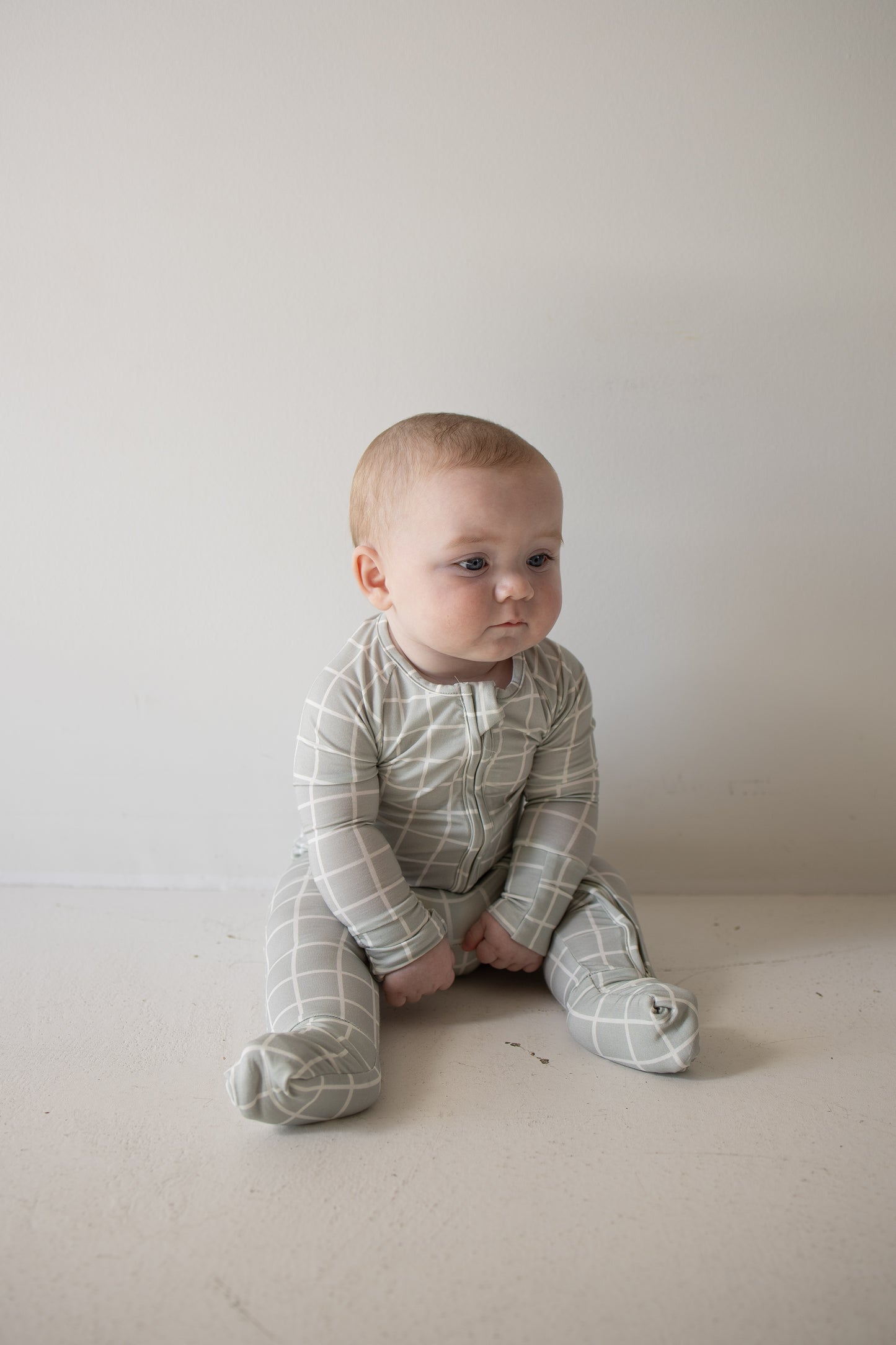 A baby in the forever french baby Bamboo Zip Pajamas | Foggy Grid sits on a light-colored floor against a plain white wall, gazing slightly to the side with a calm expression. These breathable pajamas are designed for all-day comfort, making them ideal for delicate skin.