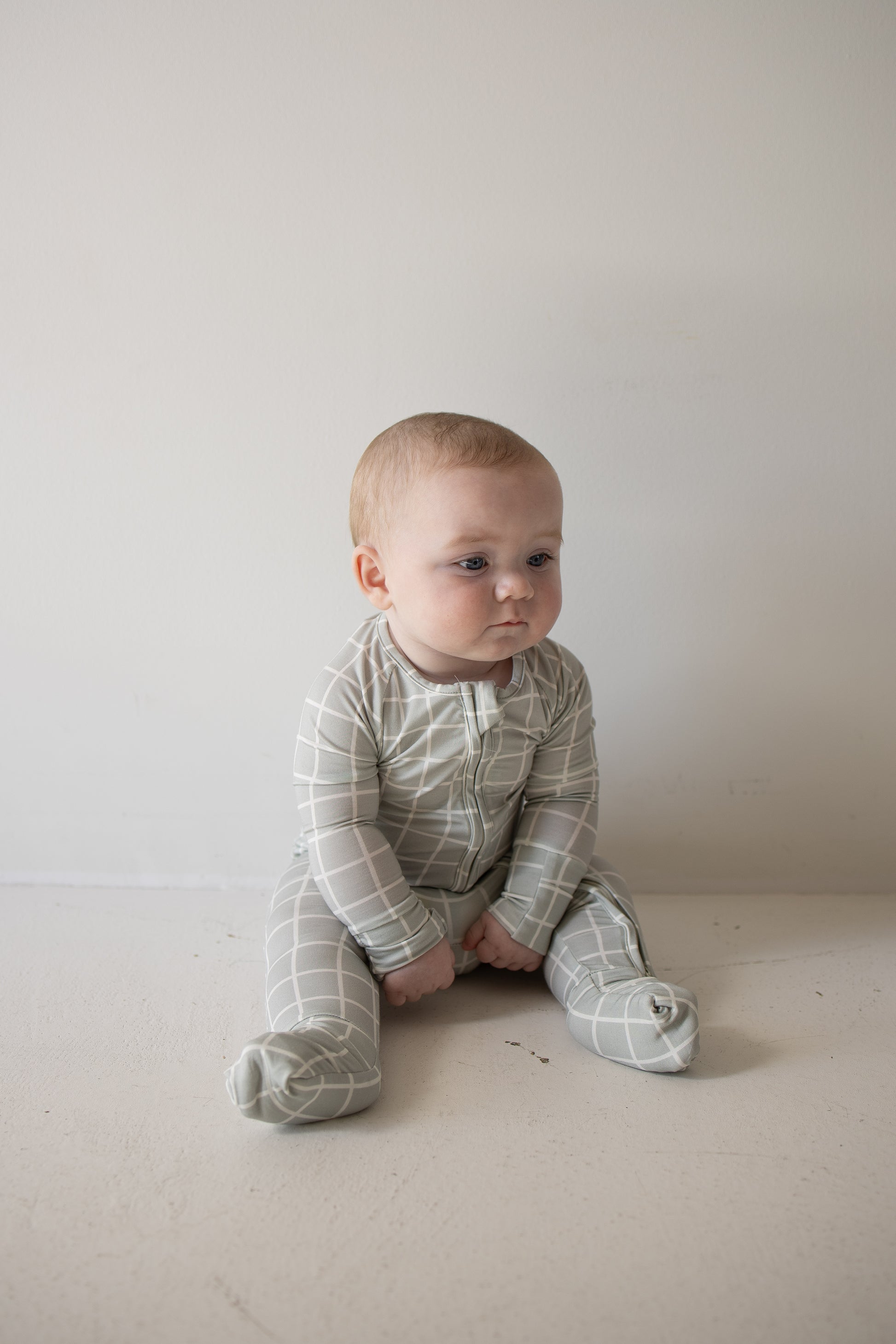 A baby in the forever french baby Bamboo Zip Pajamas | Foggy Grid sits on a light-colored floor against a plain white wall, gazing slightly to the side with a calm expression. These breathable pajamas are designed for all-day comfort, making them ideal for delicate skin.