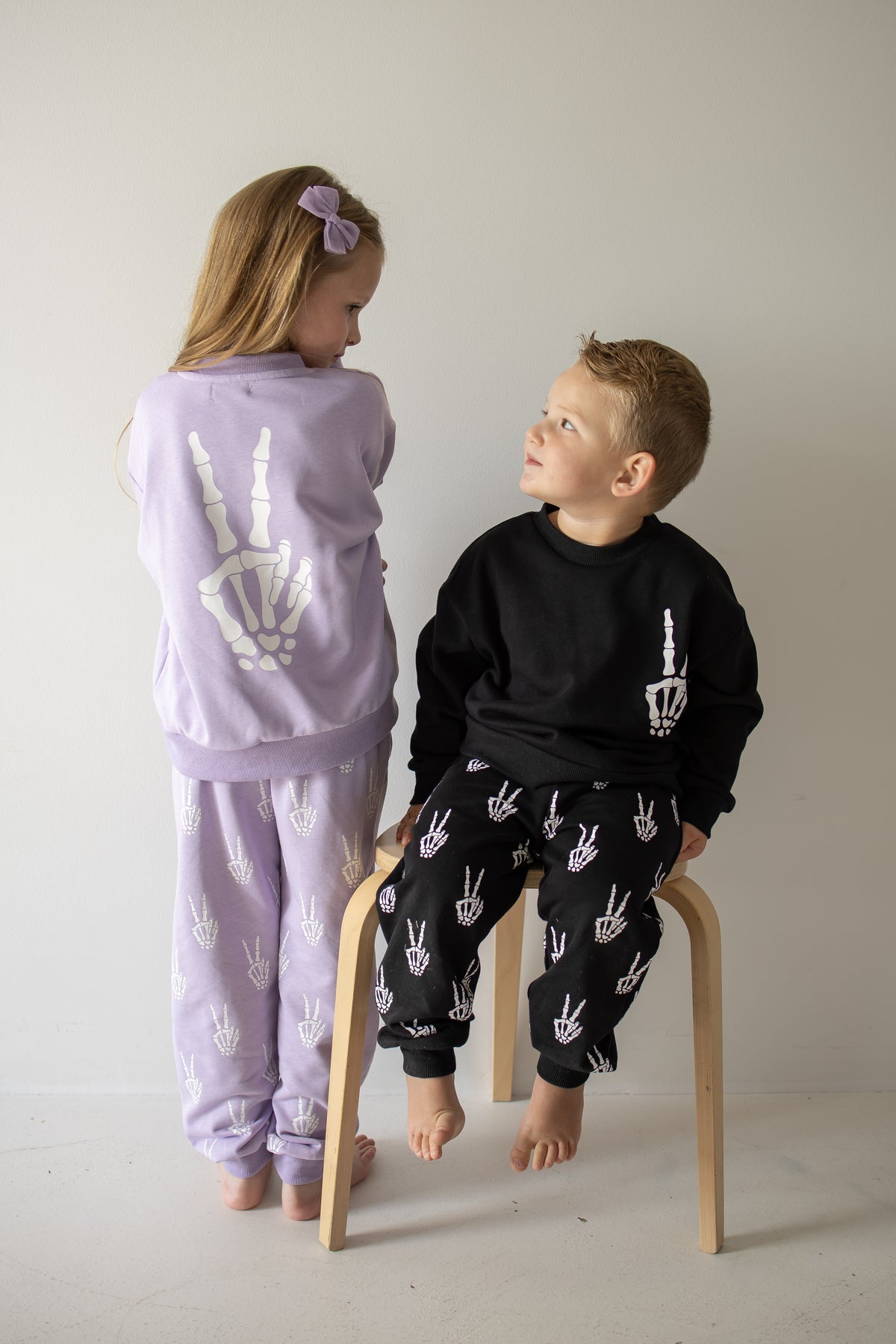 A girl wears the forever french baby Child Sweat Set | Enchanted Skelly in lavender, featuring white skeleton hand designs, while a boy sports a black version of the same set. Posing together indoors—the girl standing and the boy sitting on a wooden stool as they look at each other—they’re ready for everyday adventures.
