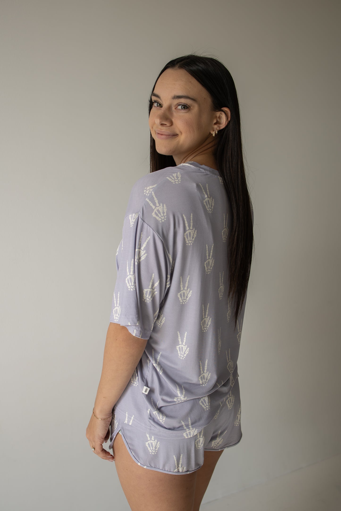 A woman with long dark hair, wearing the forever french baby Women’s Bamboo Short Set in the Enchanted Skelly print—a matching light purple pajama set with white peace sign patterns—stands sideways and smiles at the camera against a plain light background. This hypo-allergenic bamboo short set offers both comfort and style.