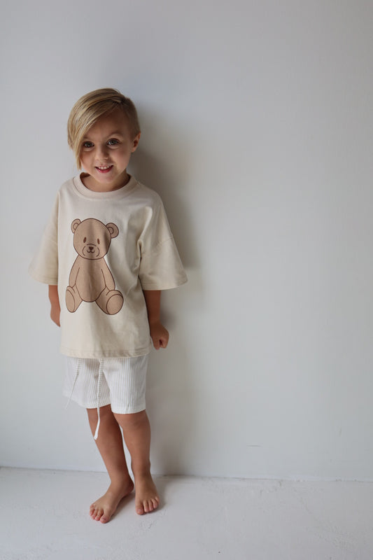 A young blonde child stands barefoot against a white wall, smiling in the forever french baby "Child Short Set | Teddy," a comfy two-piece cotton outfit with a teddy bear graphic and light shorts.