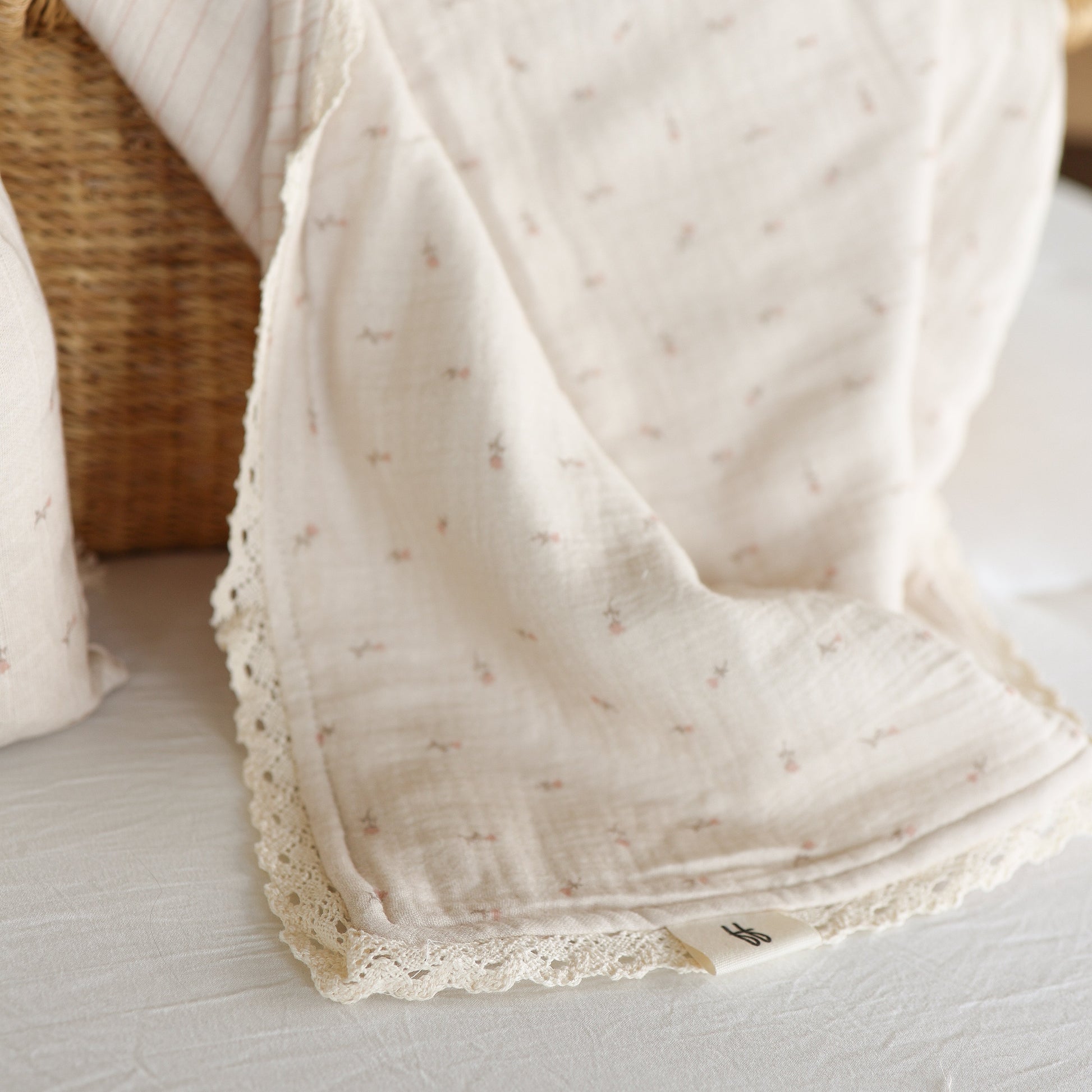 The Muslin Quilt | Lucy by forever french baby—a soft, cream-colored 100% cotton blanket with delicate lace trim and pink patterns—drapes over a wicker basket. Perfect for nursery bedding, it features a small fabric tag on the edge.