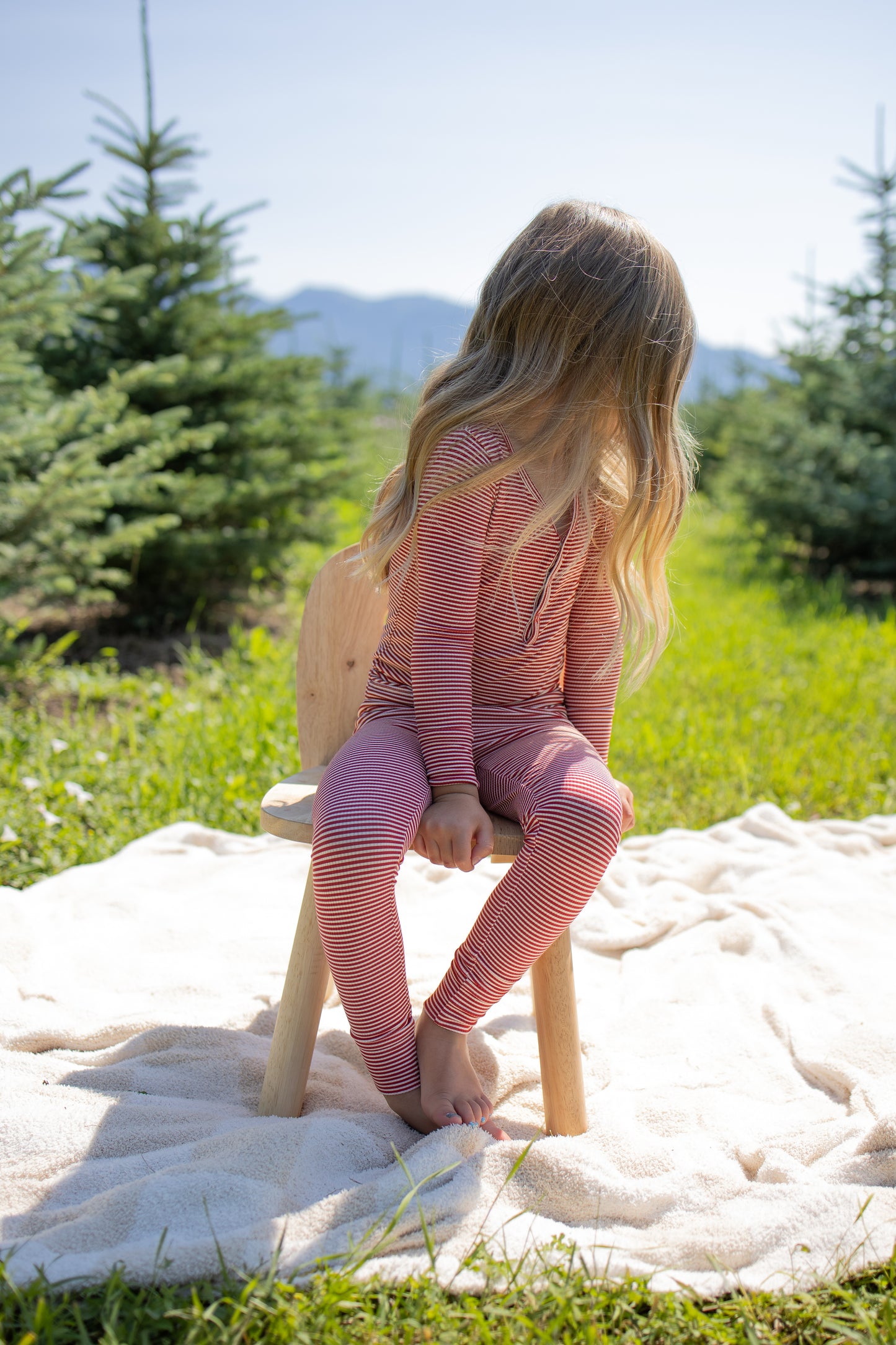 A young child with long blonde hair sits on a wooden chair outside on a blanket, wearing forever french baby’s Bamboo Two Piece Pajamas in Peppermint Stripe Ribbed, surrounded by green pine trees and sunlight.