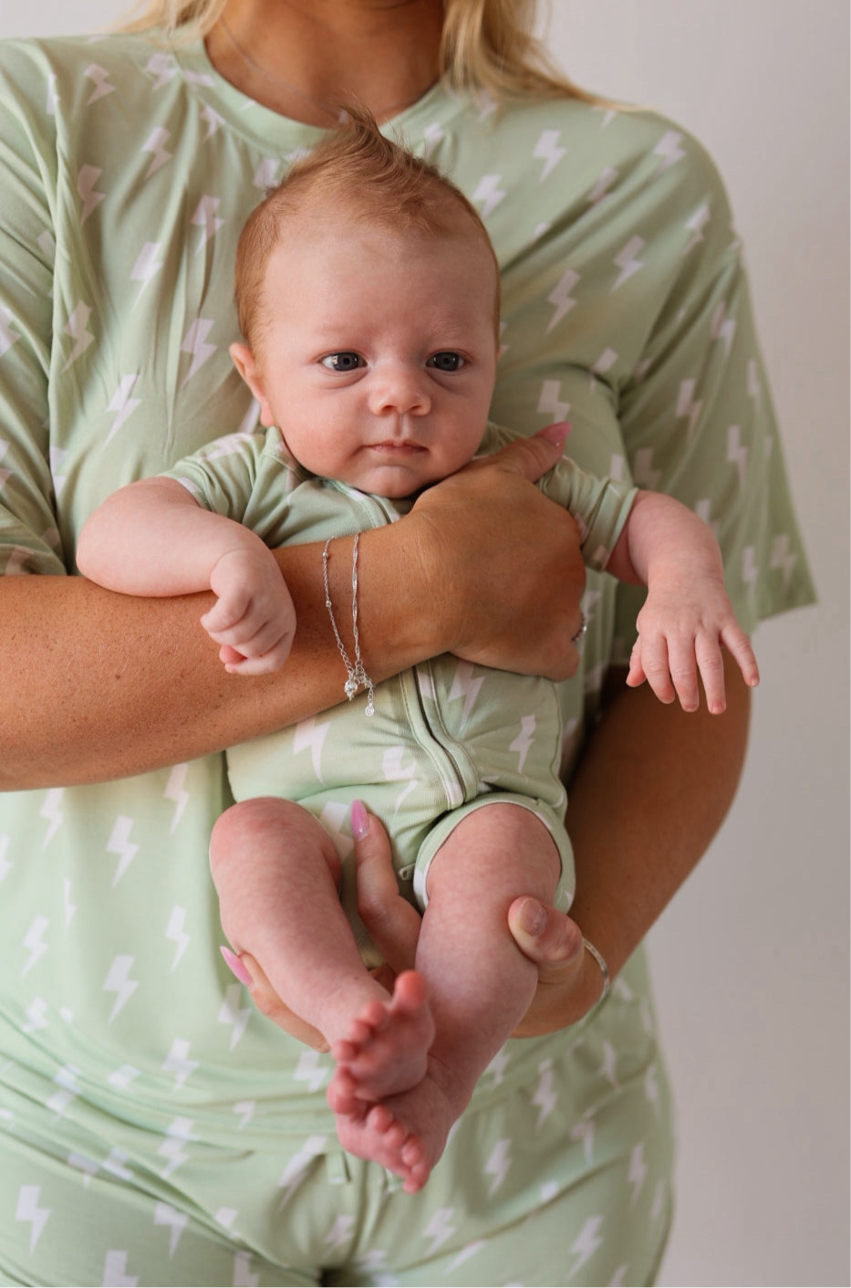 A baby with reddish hair is calmly held by an adult, both wearing forever french baby Shortie Bamboo Zip | Flash pajamas in light green with white lightning bolts, as the adult gently supports the baby.