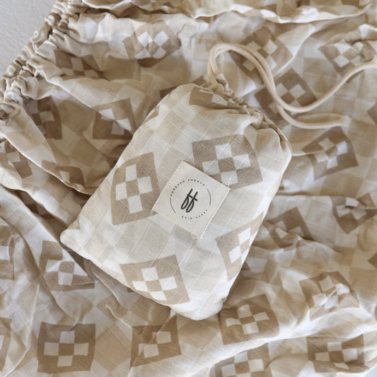 A beige geometric drawstring bag with a "bf" logo rests on matching fabric—perfect alongside the forever french baby Muslin Crib Sheet | Cottage Quilt for a cozy, neutral-toned nursery.